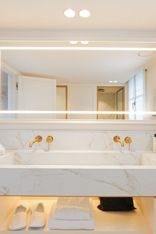 Salle de bain moderne avec double vasque en marbre blanc et robinets dorés, serviettes et chaussons rangés en dessous.