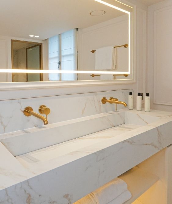 Lavabo double en marbre blanc avec robinets dorés, grand miroir éclairé, serviette blanche accrochée et produits de toilette posés sur le comptoir.