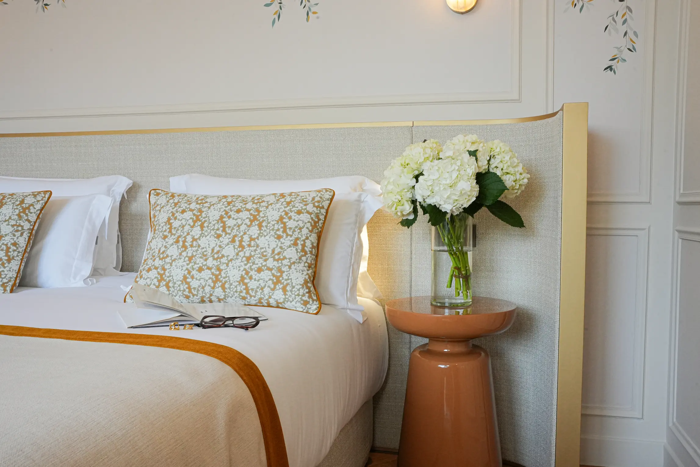 Coin lumineux d'une chambre avec un lit beige orné d'un coussin à motif floral, une table de chevet en céramique marron et un vase transparent contenant un bouquet de fleurs blanches.