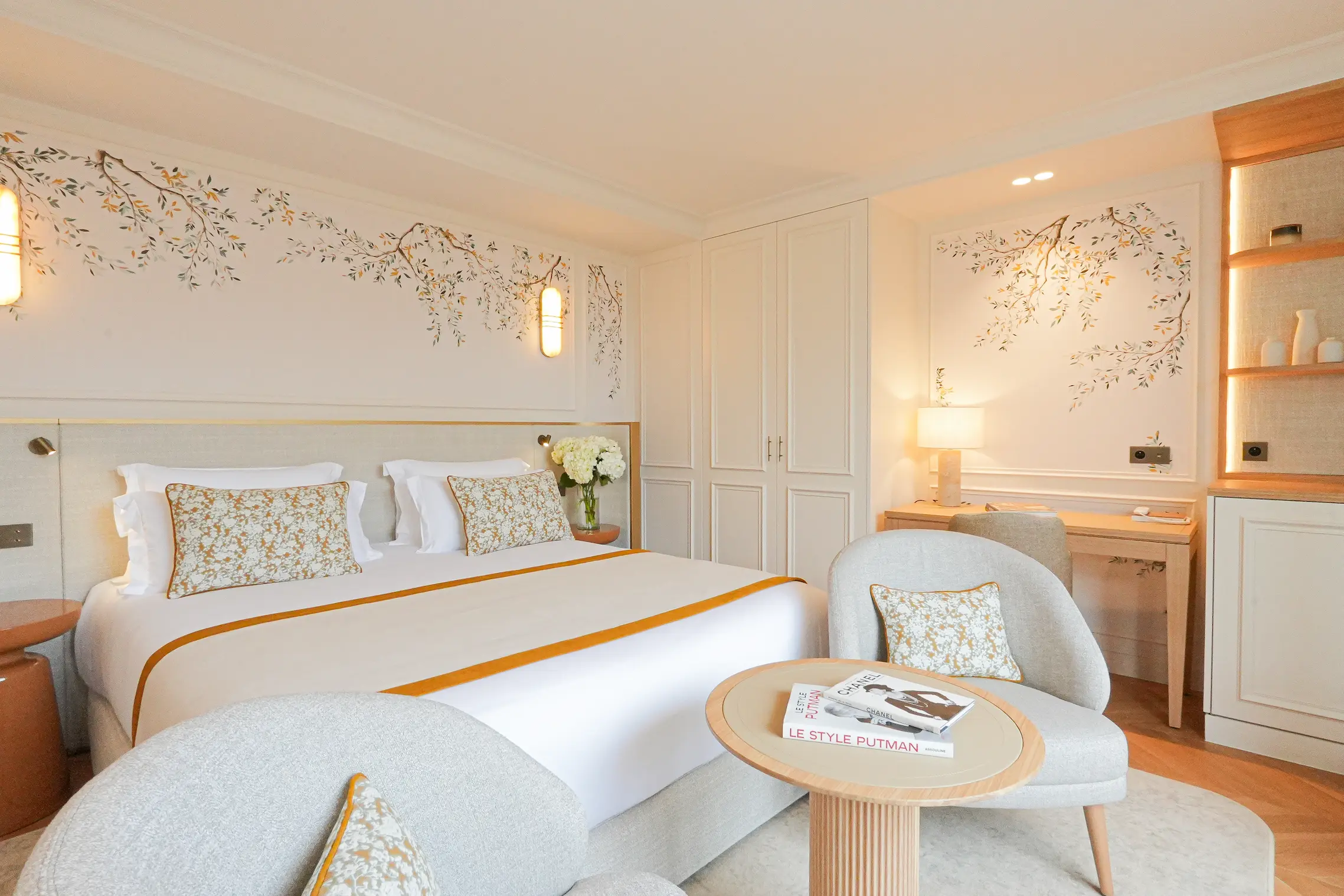 Chambre élégante avec un lit double, des fauteuils gris clairs, une table ronde en bois avec des livres, et un mur décoré de motifs floraux.