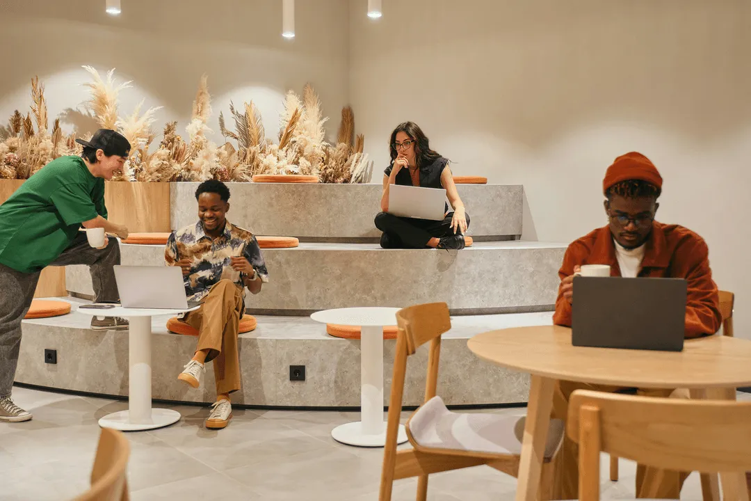 Four diverse young adults working on laptops and having coffee in a modern, cozy cafe with tiered seating and dried floral decor.