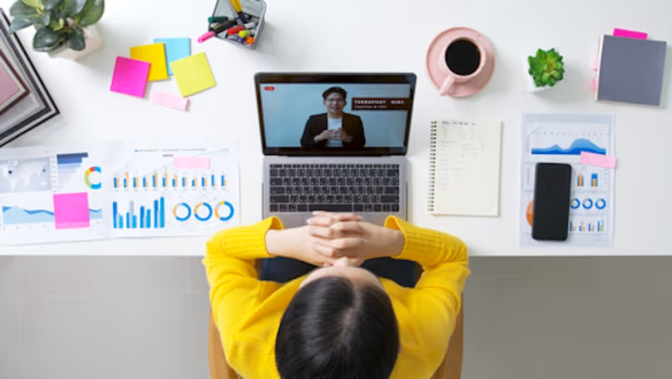 Arial view of young woman in a yellow jumper watching a video on a laptop at a white desk