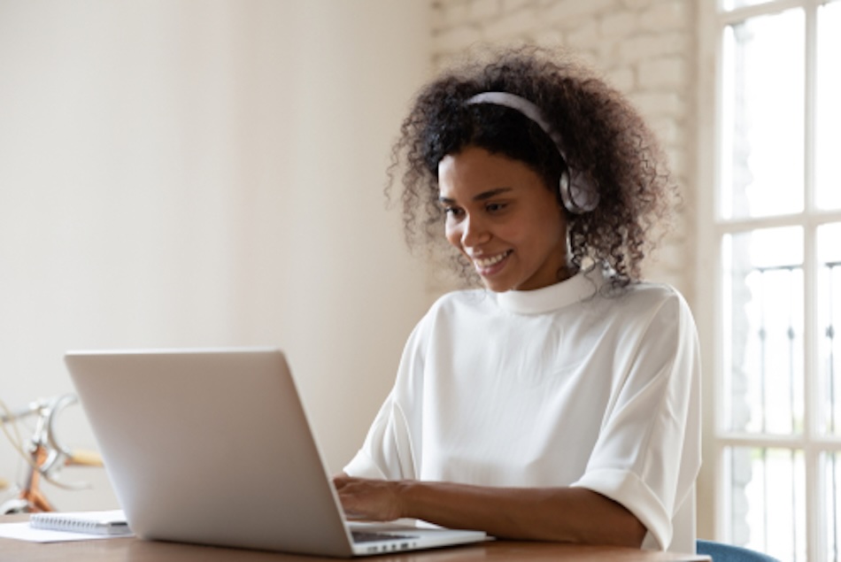 Young woman with headphones sits smiling at her laptop screen 