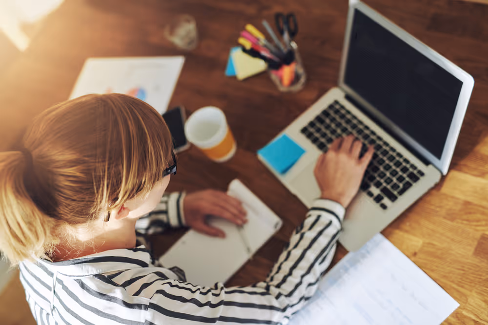 Top down view of a woman with a ponytail working at a laptop with a cup of coffee