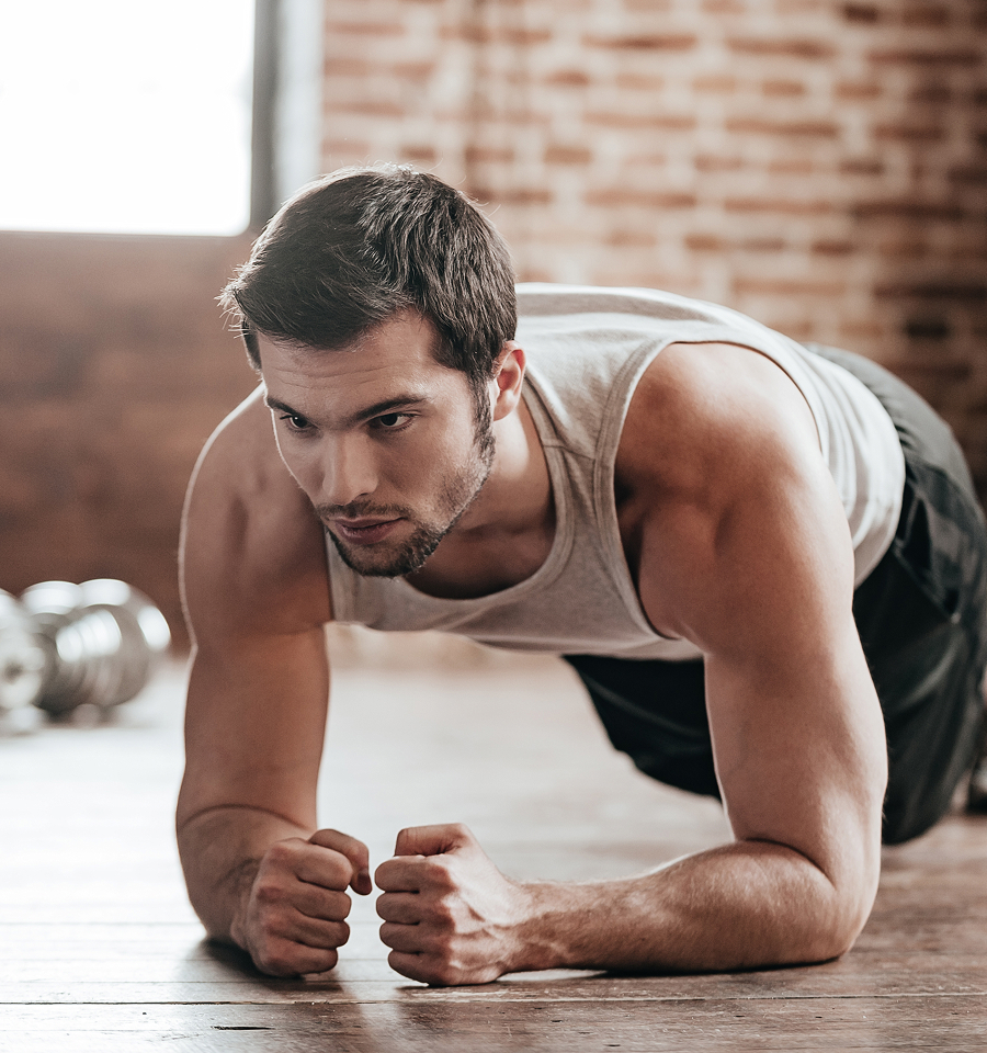 Man in a gray tank top doing a plank exercise on a wooden floor in a gym with brick walls.