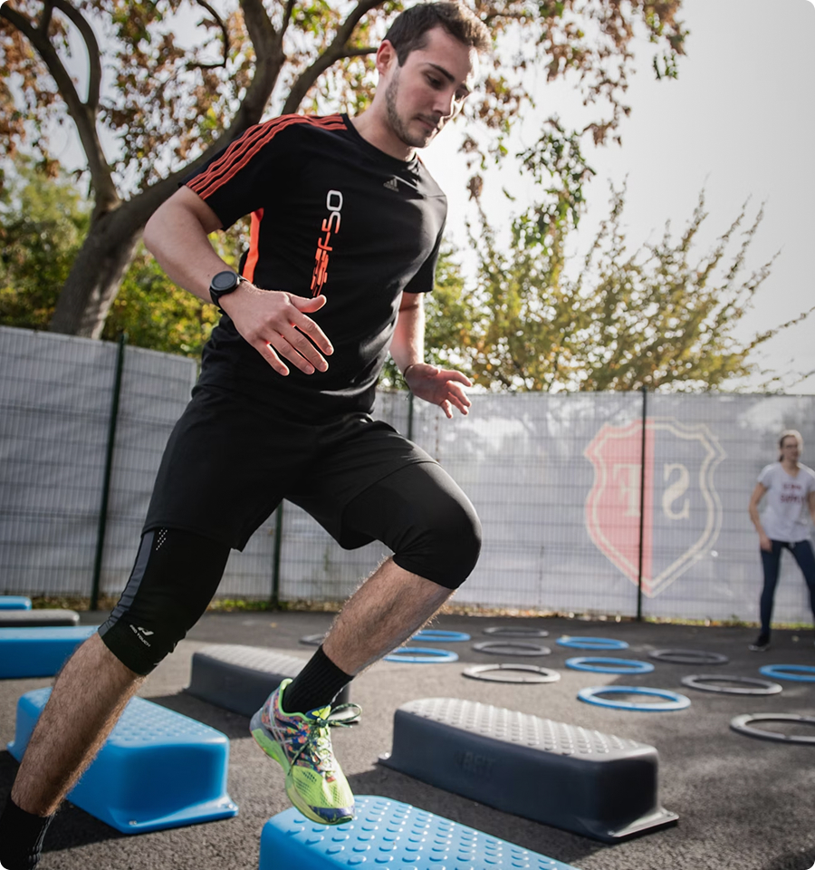 Man in black sportswear and neon running shoes exercising outdoors on blue stepping platforms in an agility drill.