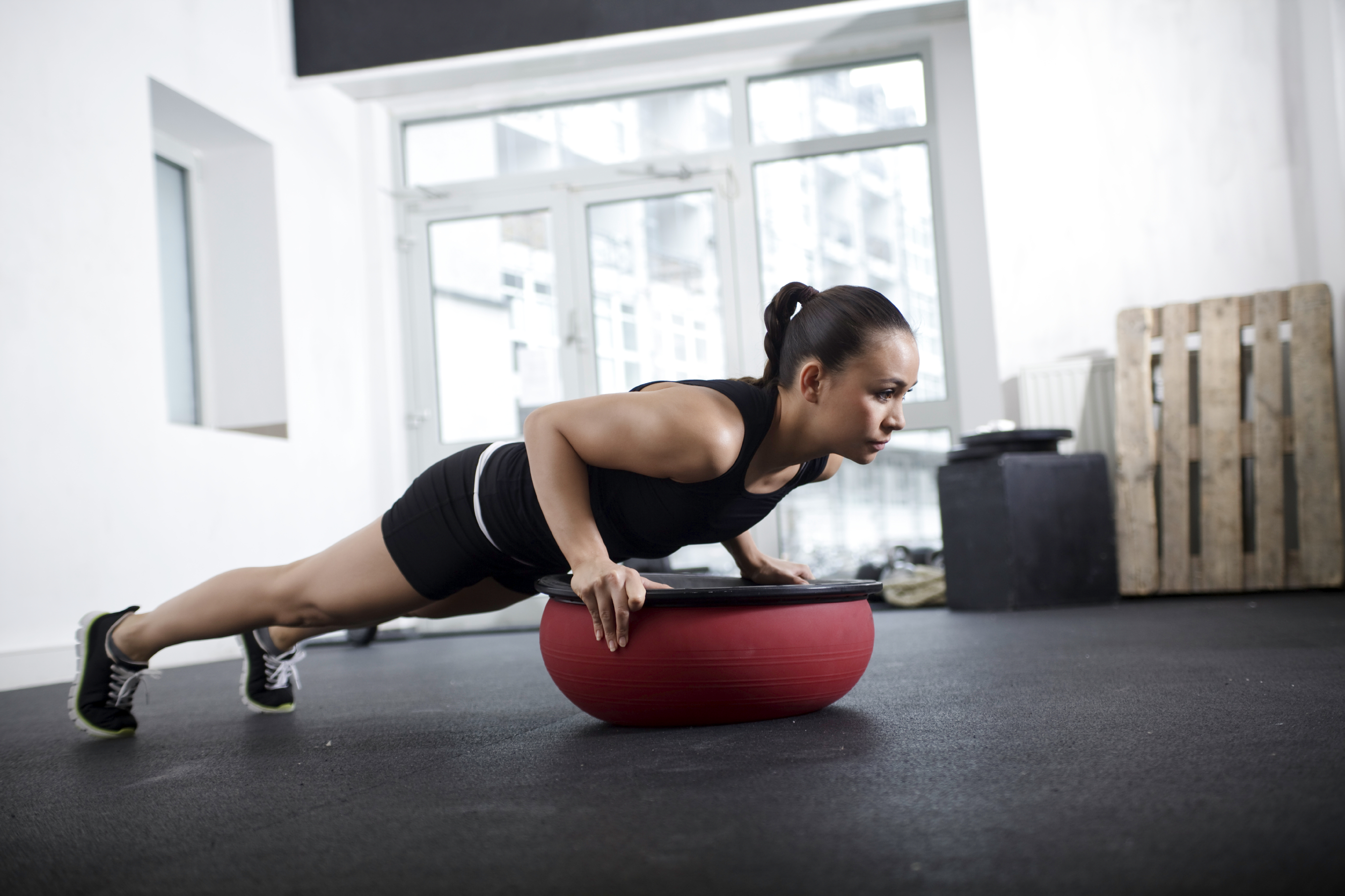 Woman performing push-up exercise on a red balance trainer in a gym with large windows.