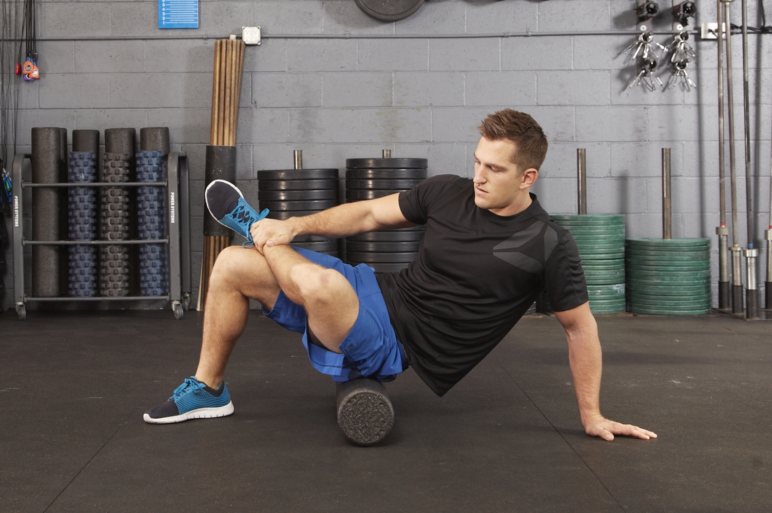Man in black shirt and blue shorts using a foam roller under his back while holding his ankle in a gym.