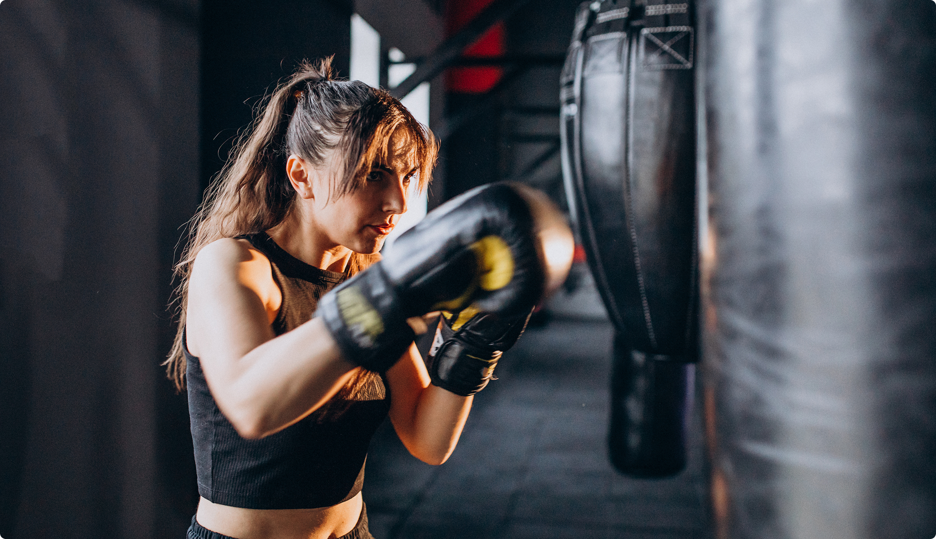 Woman with ponytail wearing black sportswear and boxing gloves punching a black punching bag in a gym.