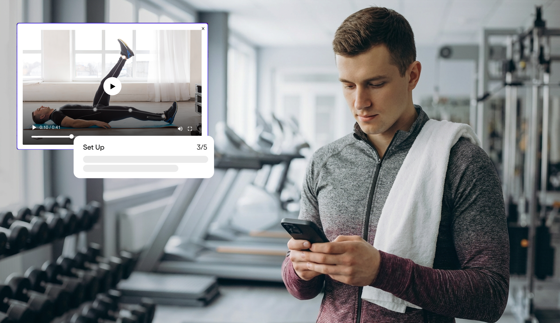 Man in gym wearing a gray and maroon jacket with a white towel over his shoulder, looking at his smartphone with treadmills and weights in the background, alongside an exercise video overlay showing a man doing leg lifts on a mat.