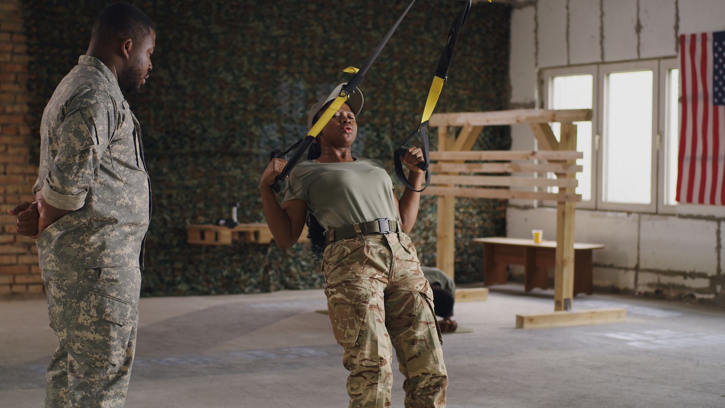 Female soldier in camouflage pants and green t-shirt exercising with suspension straps while a male soldier watches.