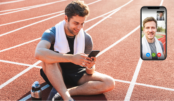Man sitting on a running track with a towel around his neck, holding a smartphone and video calling another man displayed on the phone screen.
