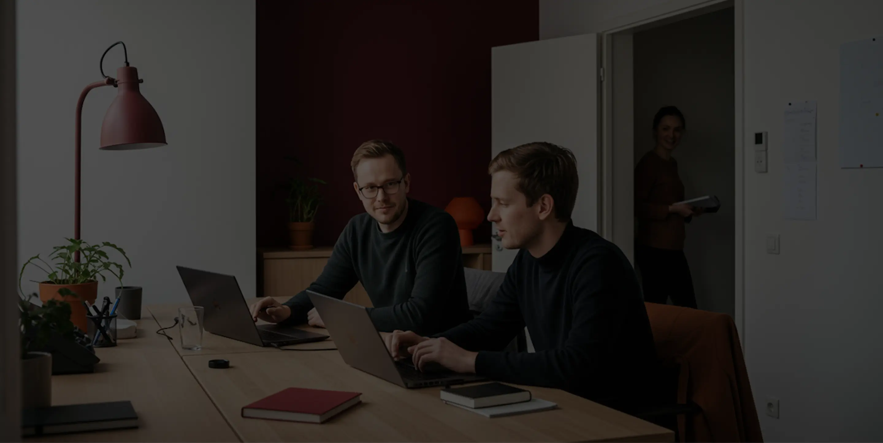 Two men working on laptops at a wooden table in an office, with a woman holding a notebook standing in a doorway behind them.