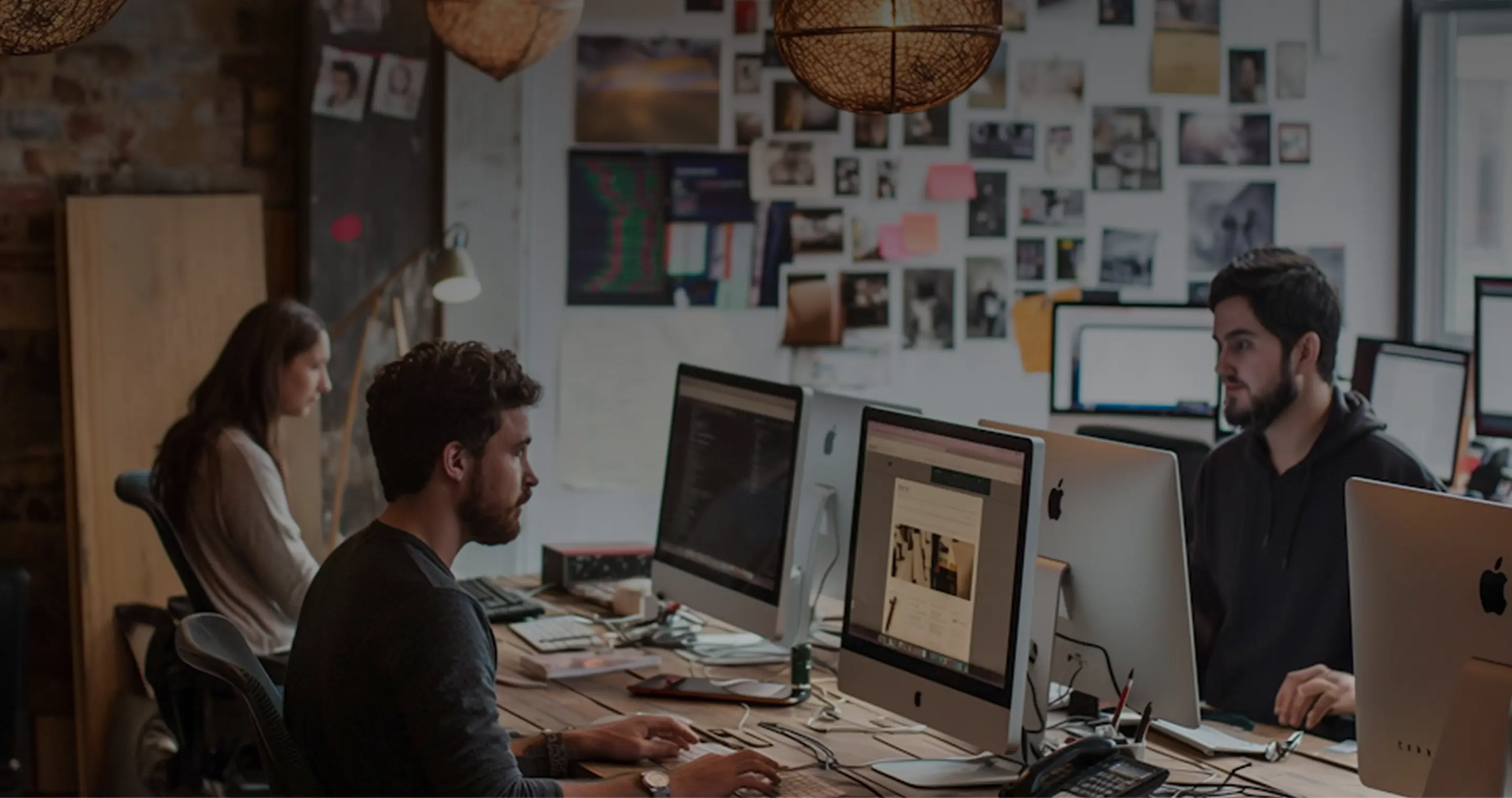 Three people working at a shared desk with multiple Apple iMac computers in a modern office with a photo-covered wall.