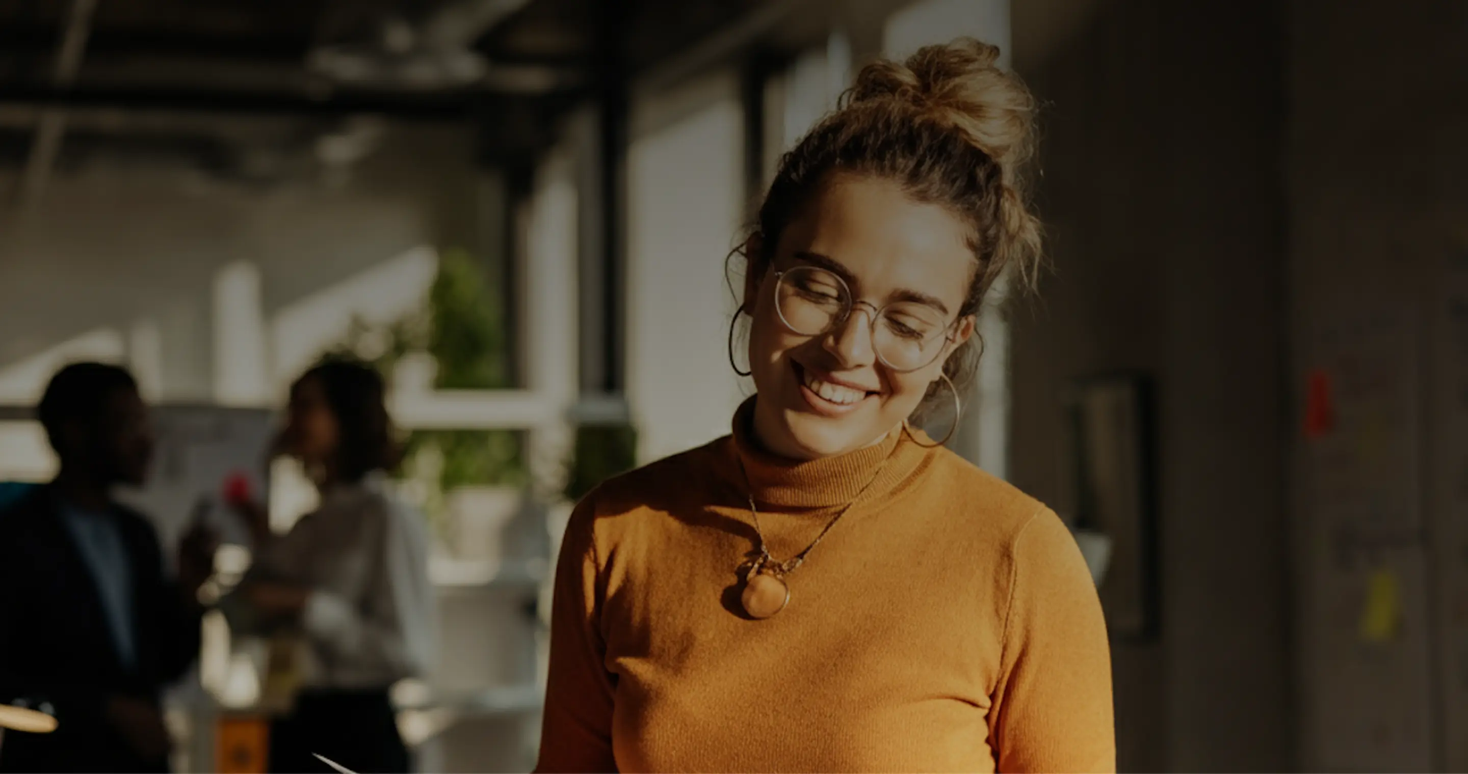 Smiling woman with glasses and hair in a bun, wearing an orange turtleneck sweater and necklace in a bright office with blurred people in the background.