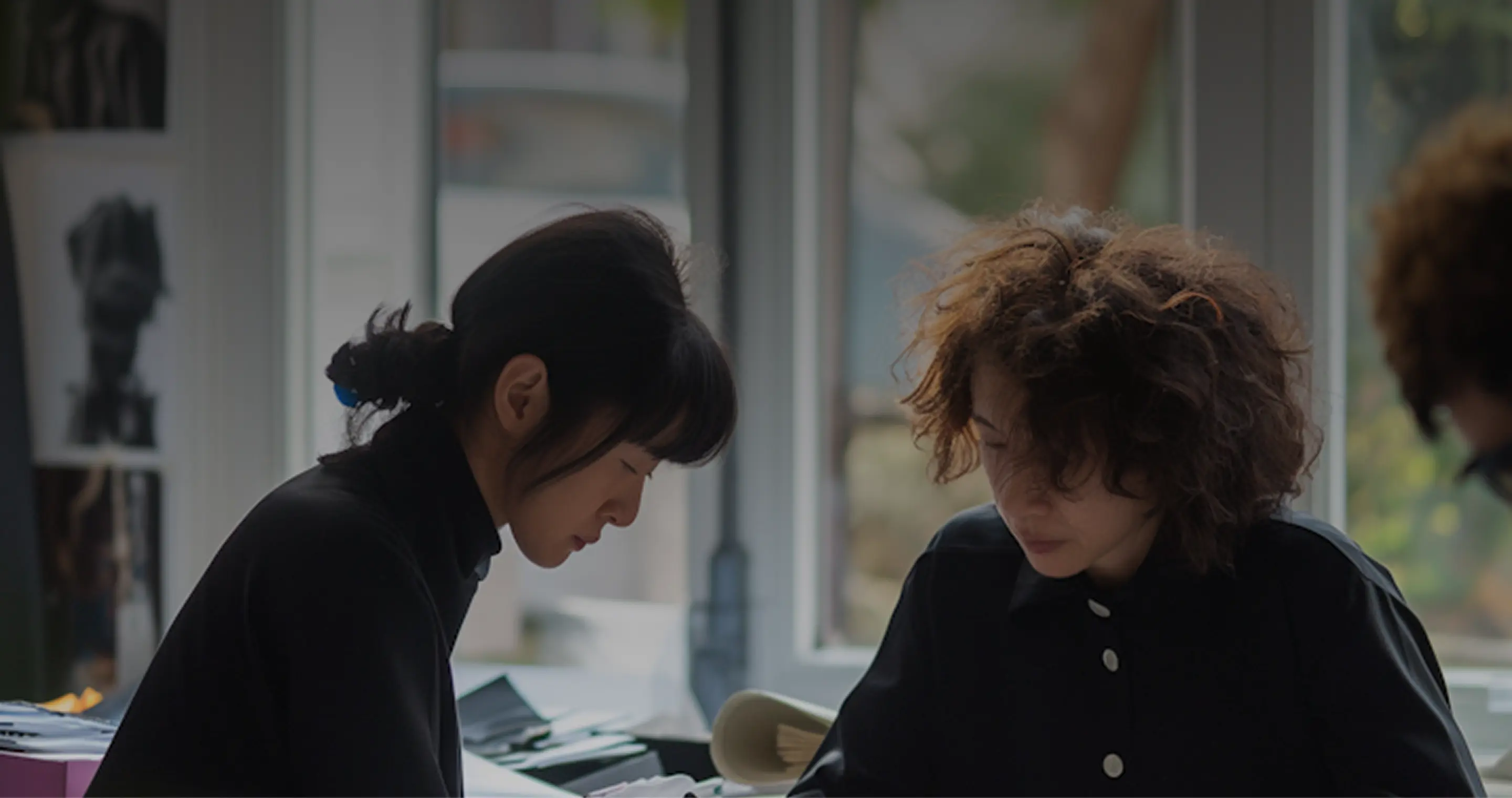 Two women focused on their work at a desk by a window in a softly lit room.