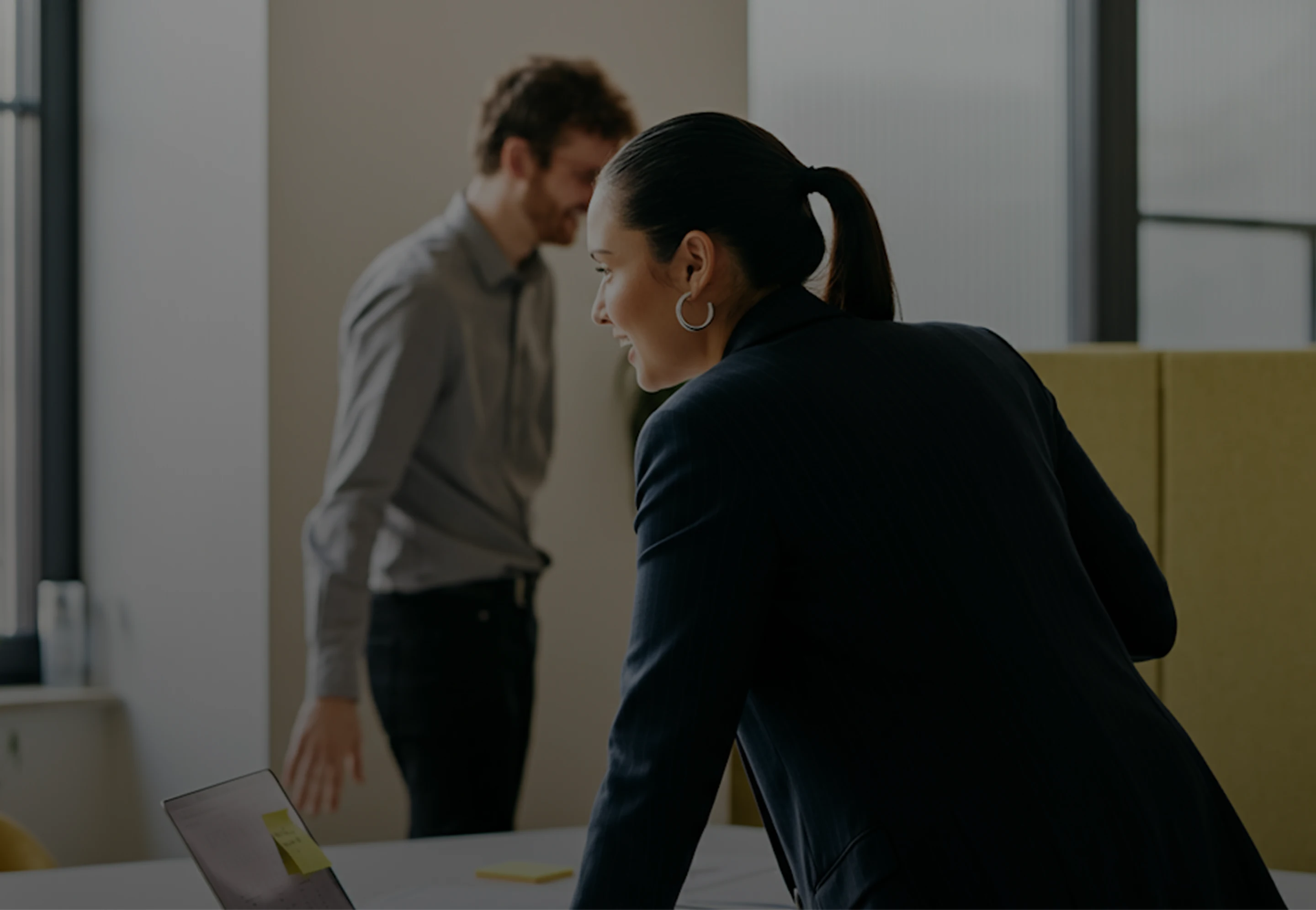 Smiling woman in business attire leaning over a table with a laptop, with a man standing and blurred in the background.