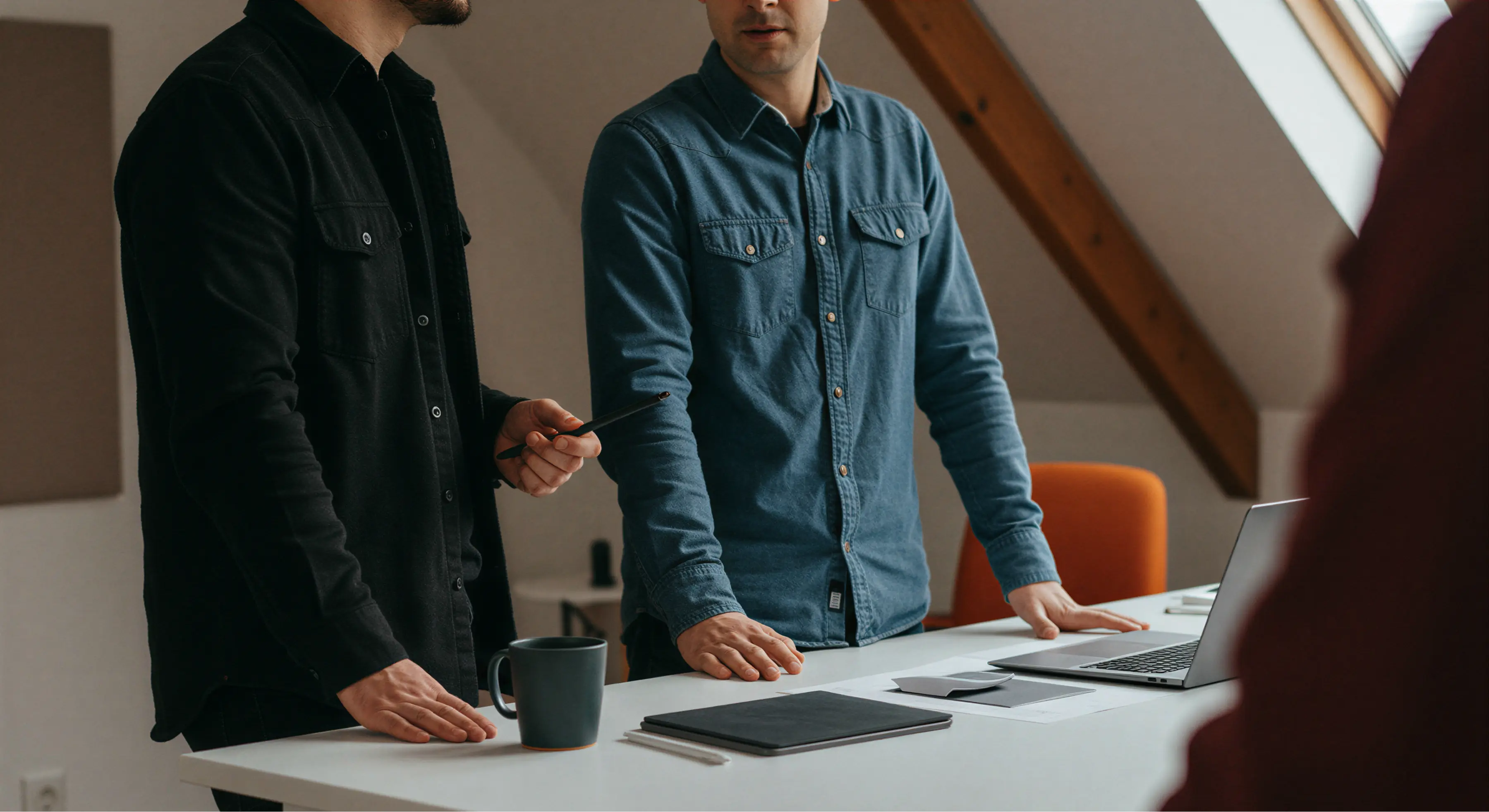 Two men standing and discussing over a white table with laptop, documents, and a coffee mug in an office.
