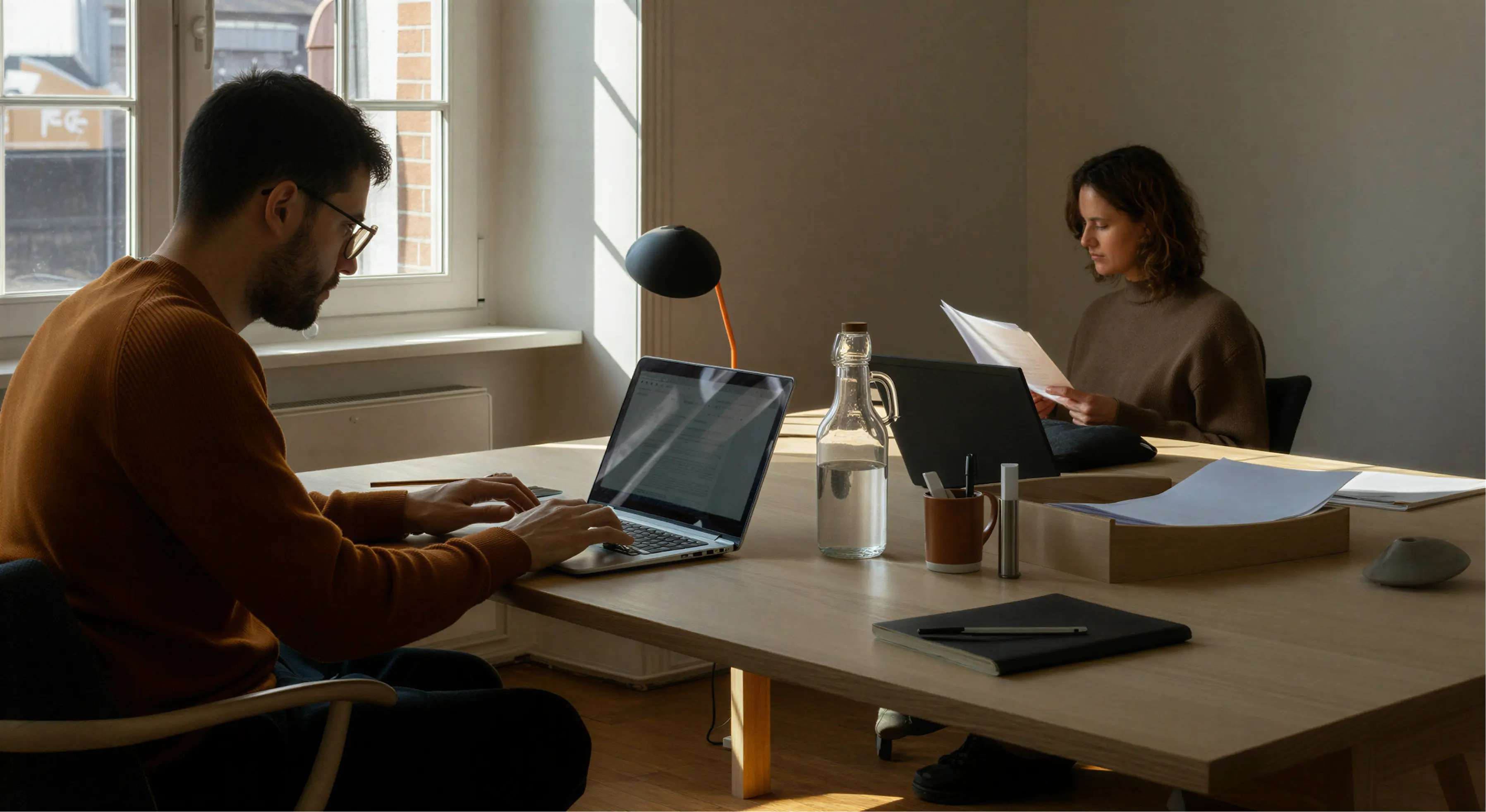Two people working quietly at a wooden table with laptops and papers in a sunlit office.