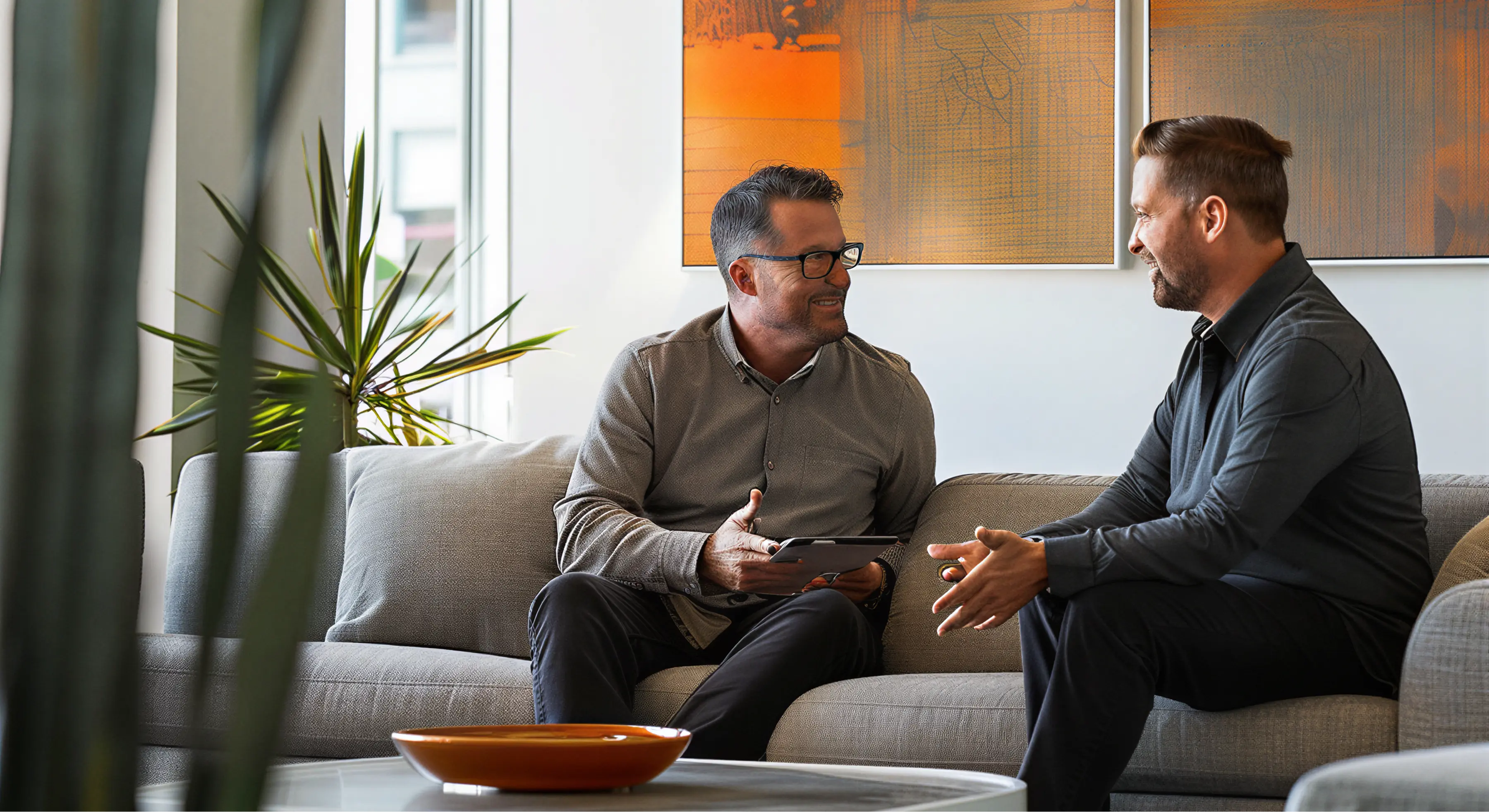 Two men sitting on a gray sofa in a modern office, engaged in a friendly conversation with one holding a tablet.
