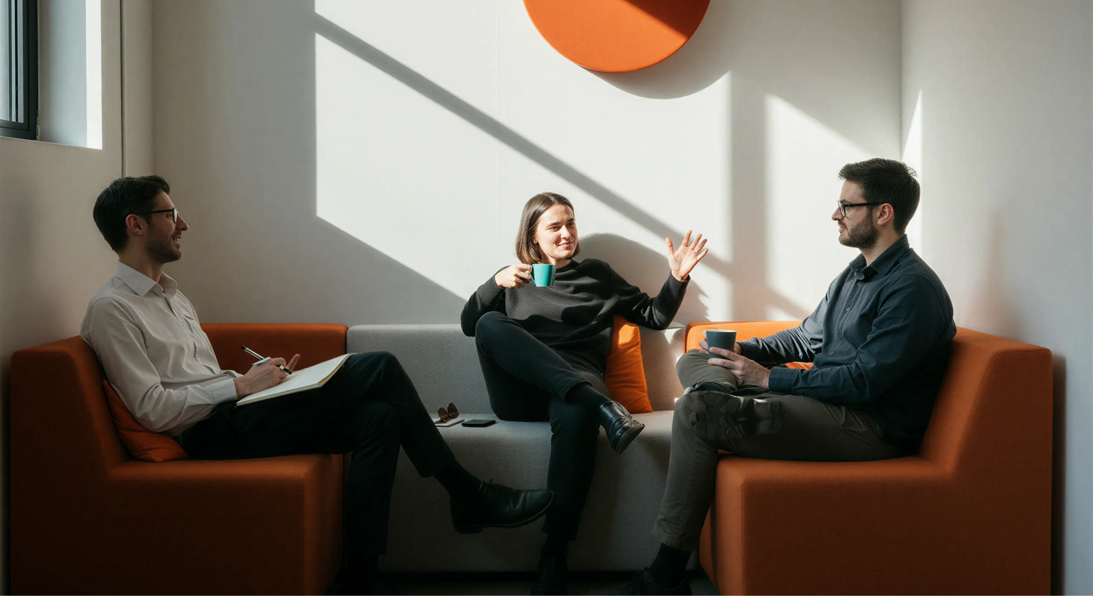 Three people having a discussion while sitting on modern orange and gray sofas in a sunlit room, two holding coffee mugs and one taking notes.