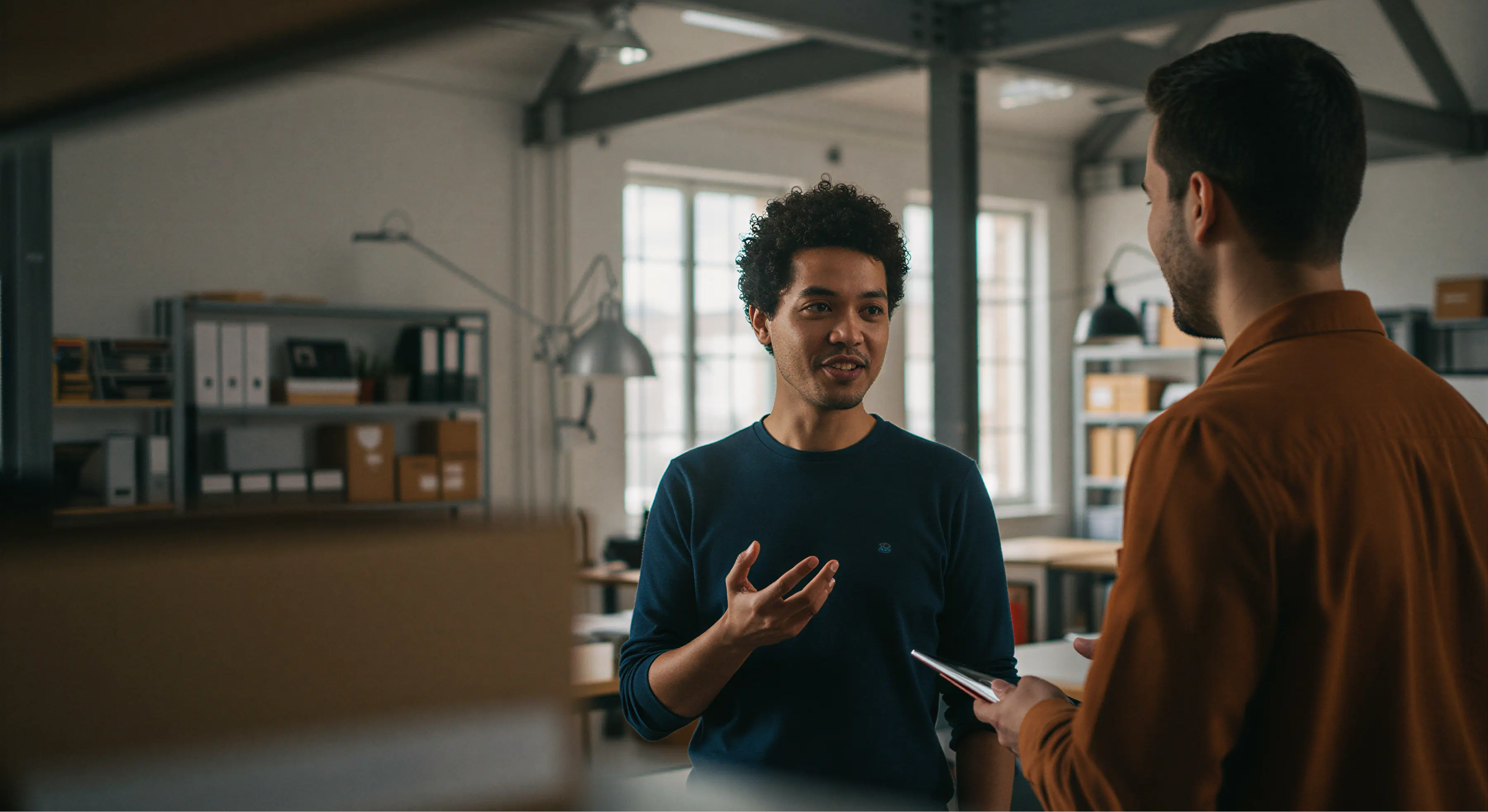 Two men having a casual discussion in a modern office with shelves and large windows in the background.