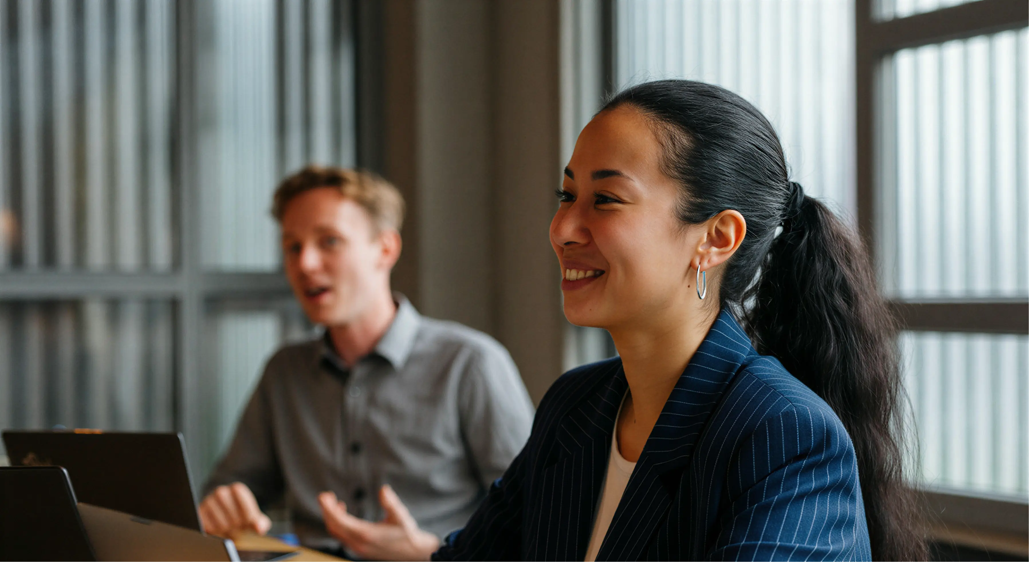 Smiling woman with a ponytail and hoop earrings listens while a man speaks in a modern office setting.