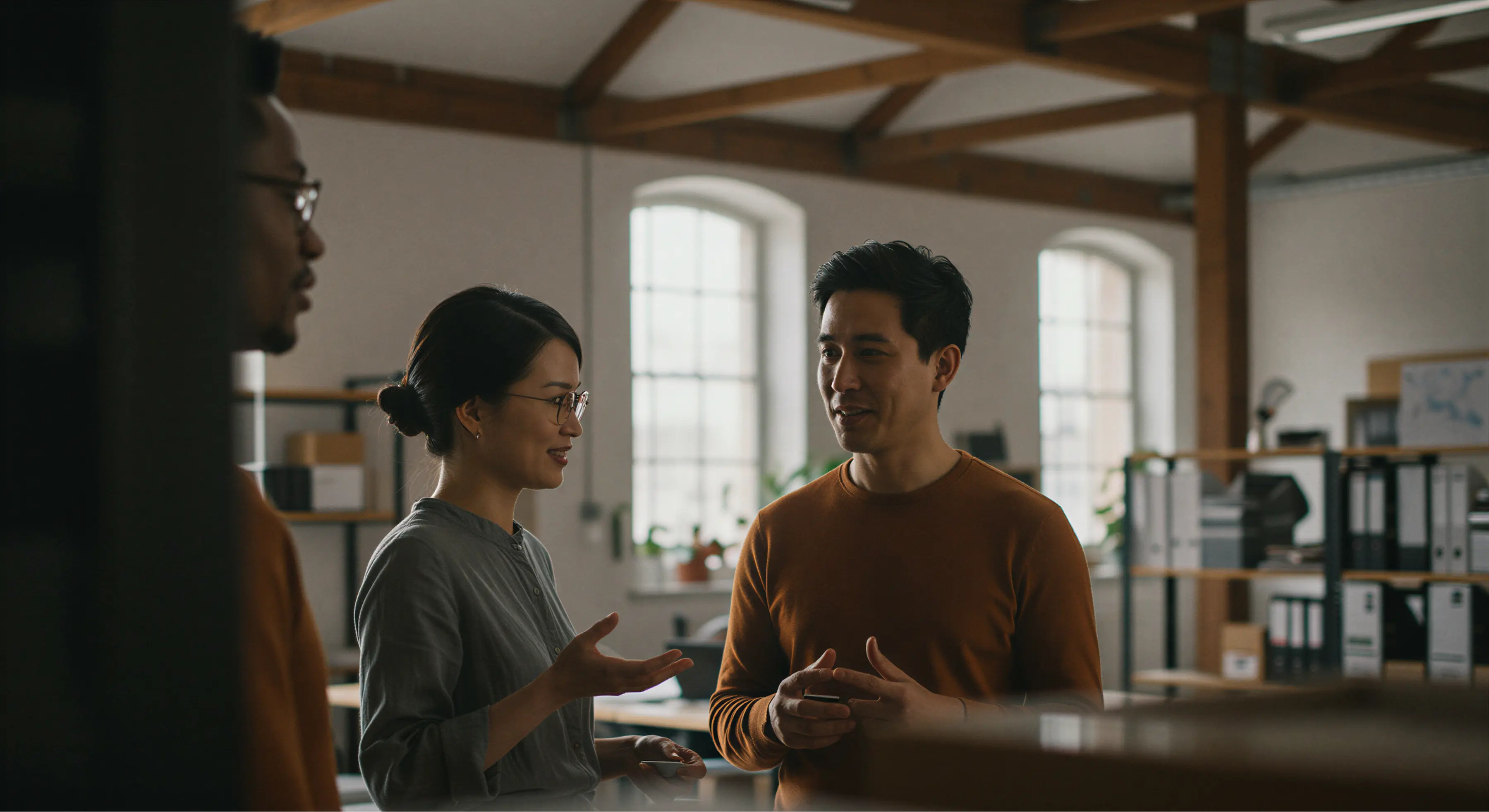 Three colleagues having a discussion in a modern office with large windows and shelves with binders in the background.