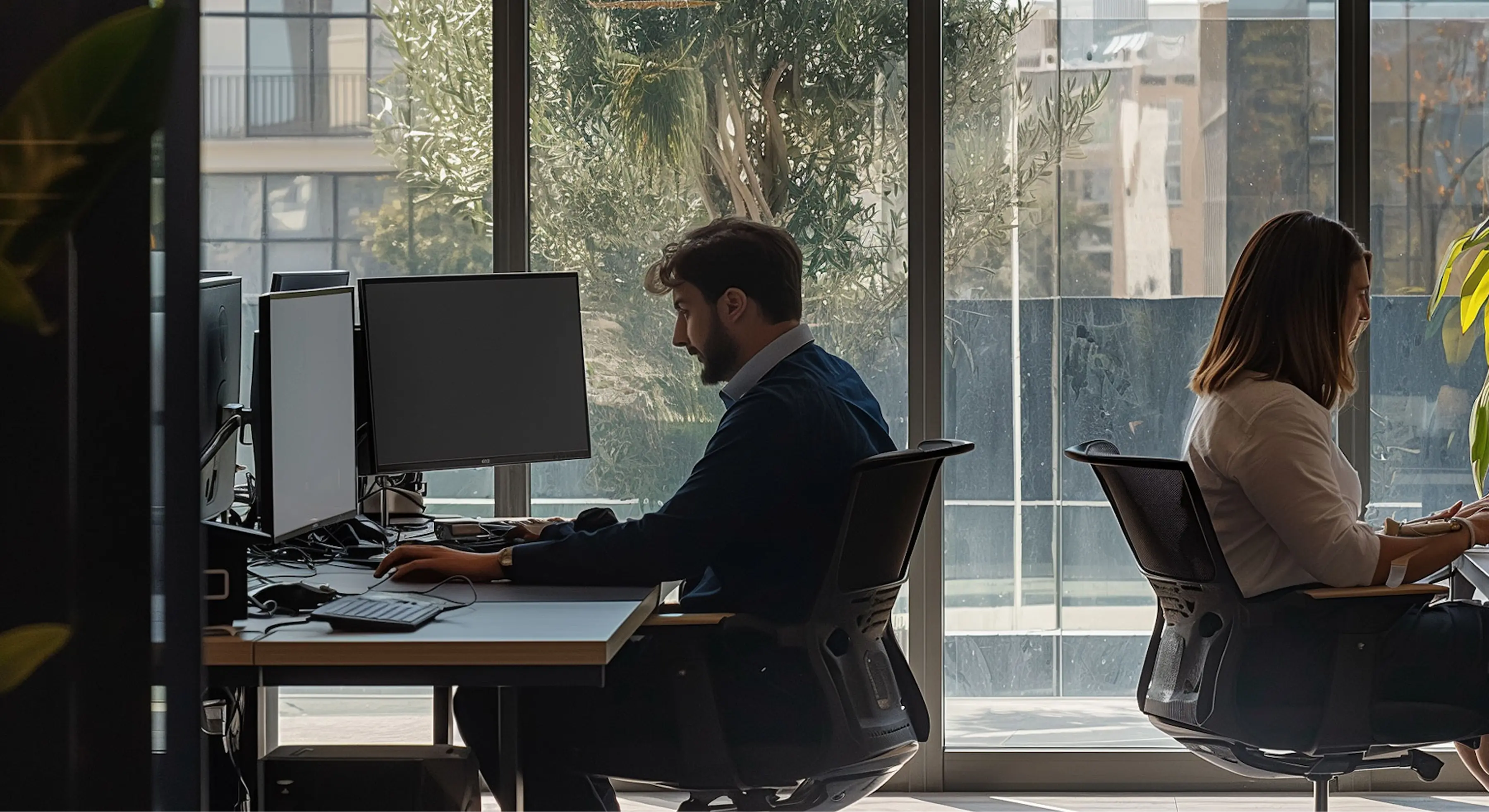 Two people working on computers at desks in a modern office with large windows and natural light.