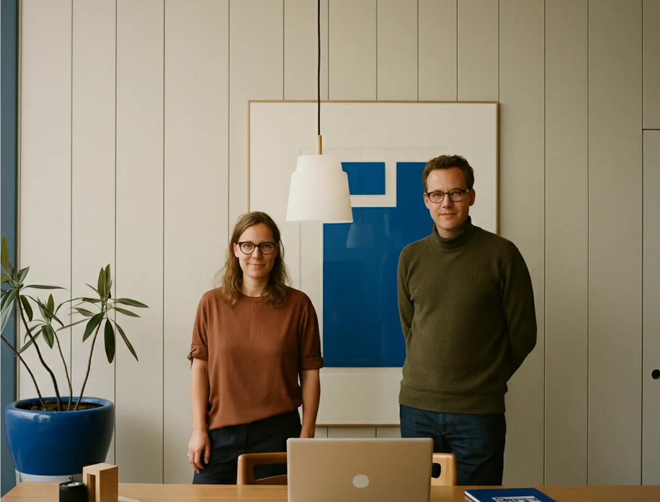 A woman and a man wearing glasses standing behind a wooden desk with a laptop, a blue potted plant to the side, and a blue abstract framed artwork on the wall behind them.