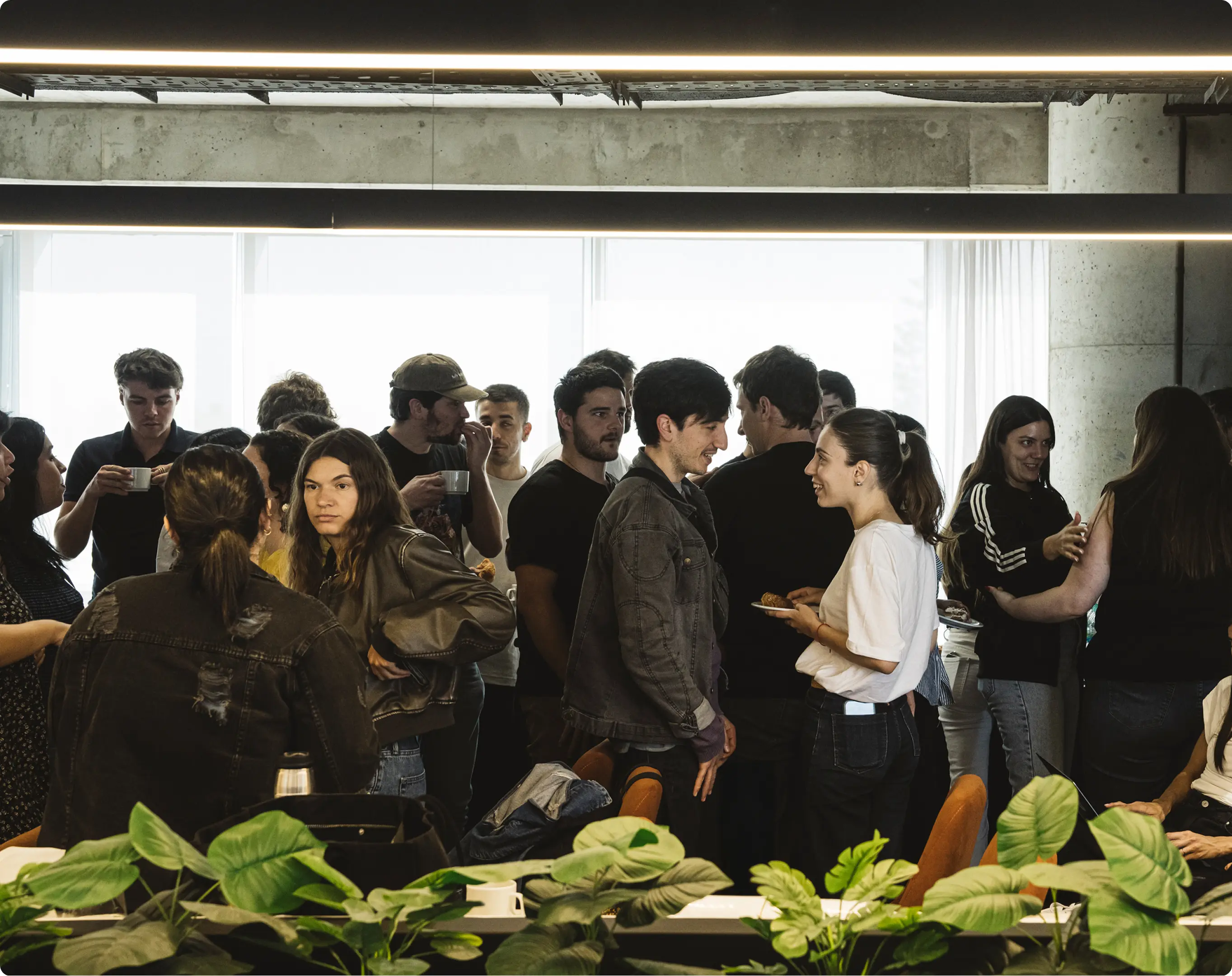 Group of young people socializing indoors near a window with greenery in the foreground.