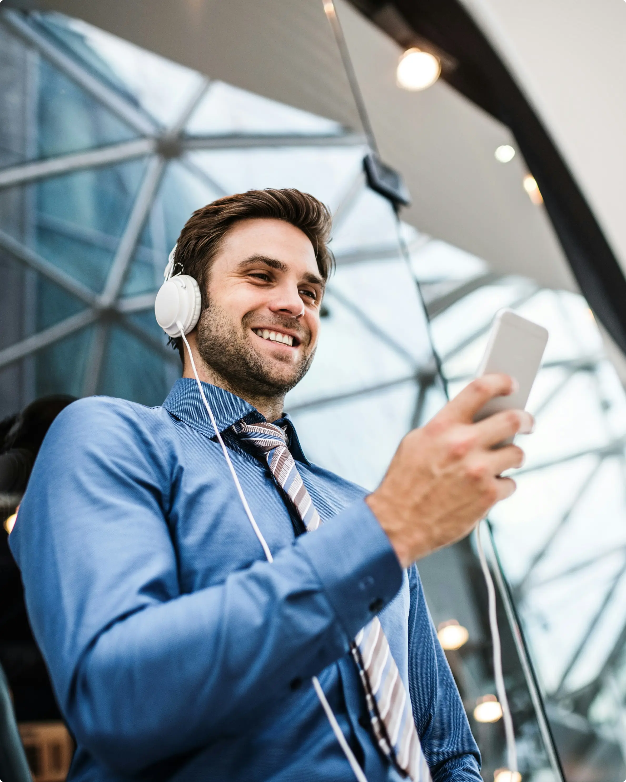 Smiling man in a blue dress shirt and striped tie listening to headphones and looking at his smartphone.