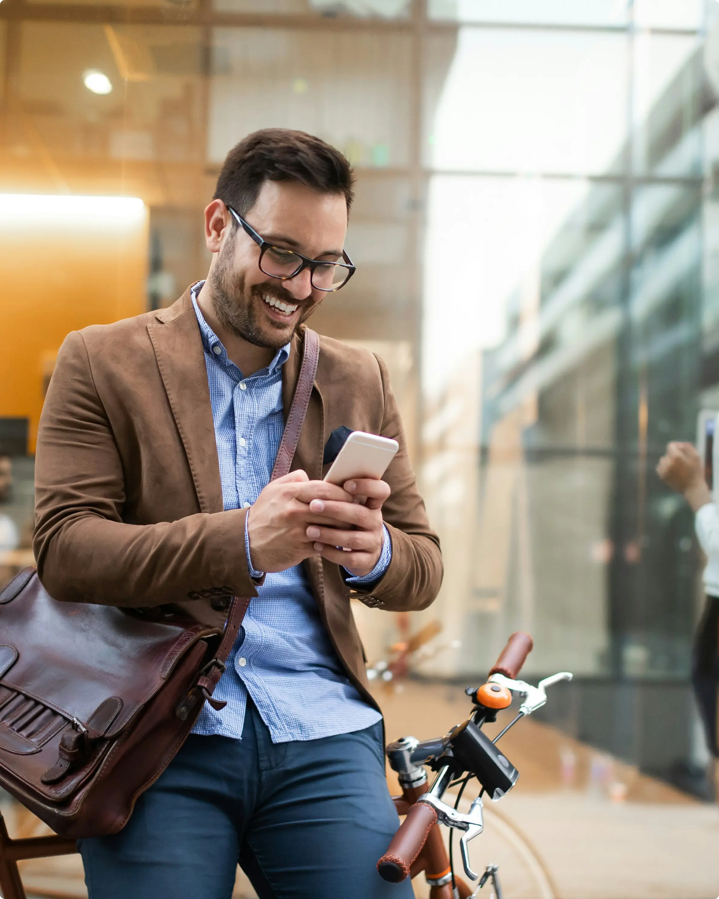 Smiling man in glasses, brown blazer, and blue shirt using a smartphone while standing next to a bicycle with a brown leather satchel.