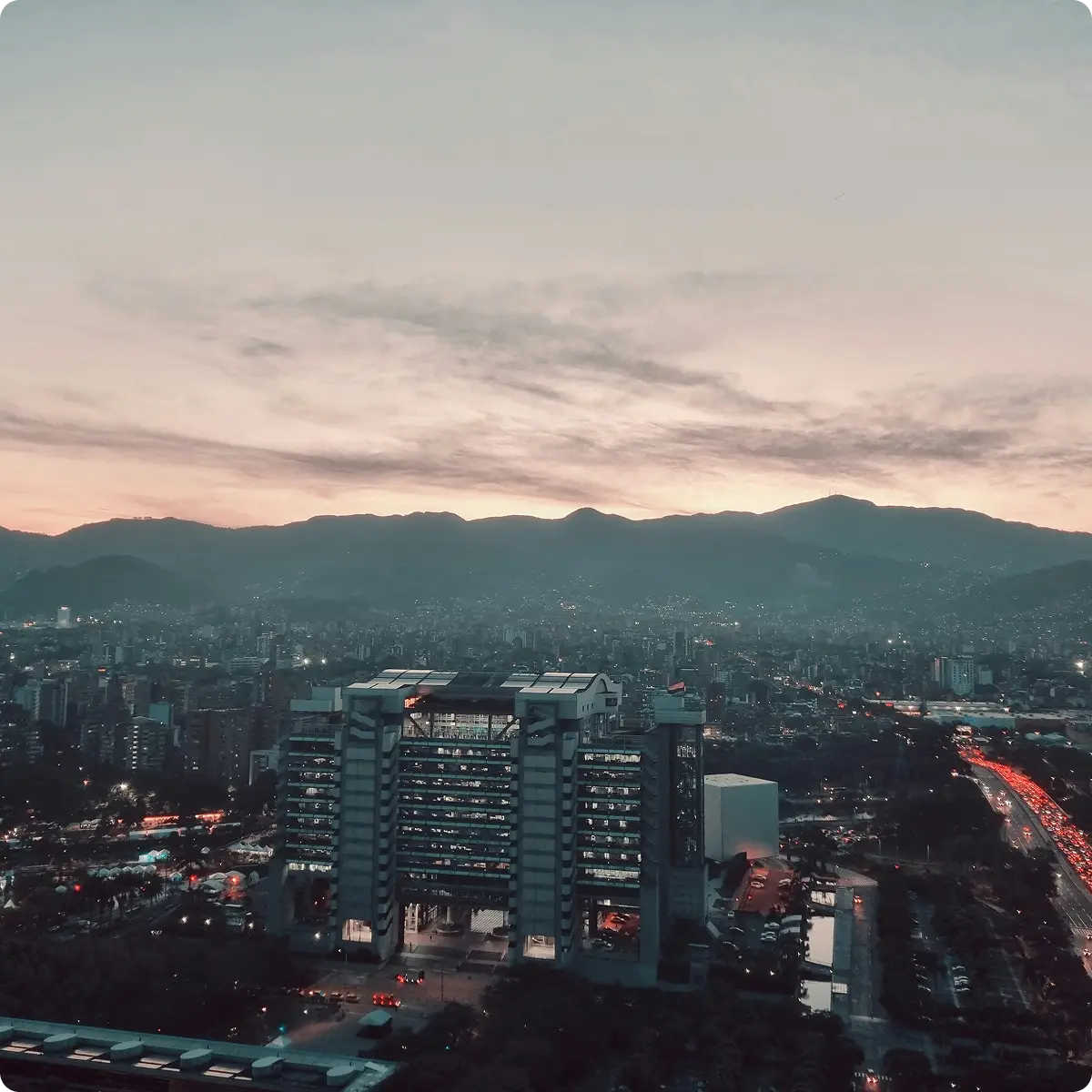 Cityscape at dusk with a large modern building in the foreground and mountains in the background under a cloudy sky.