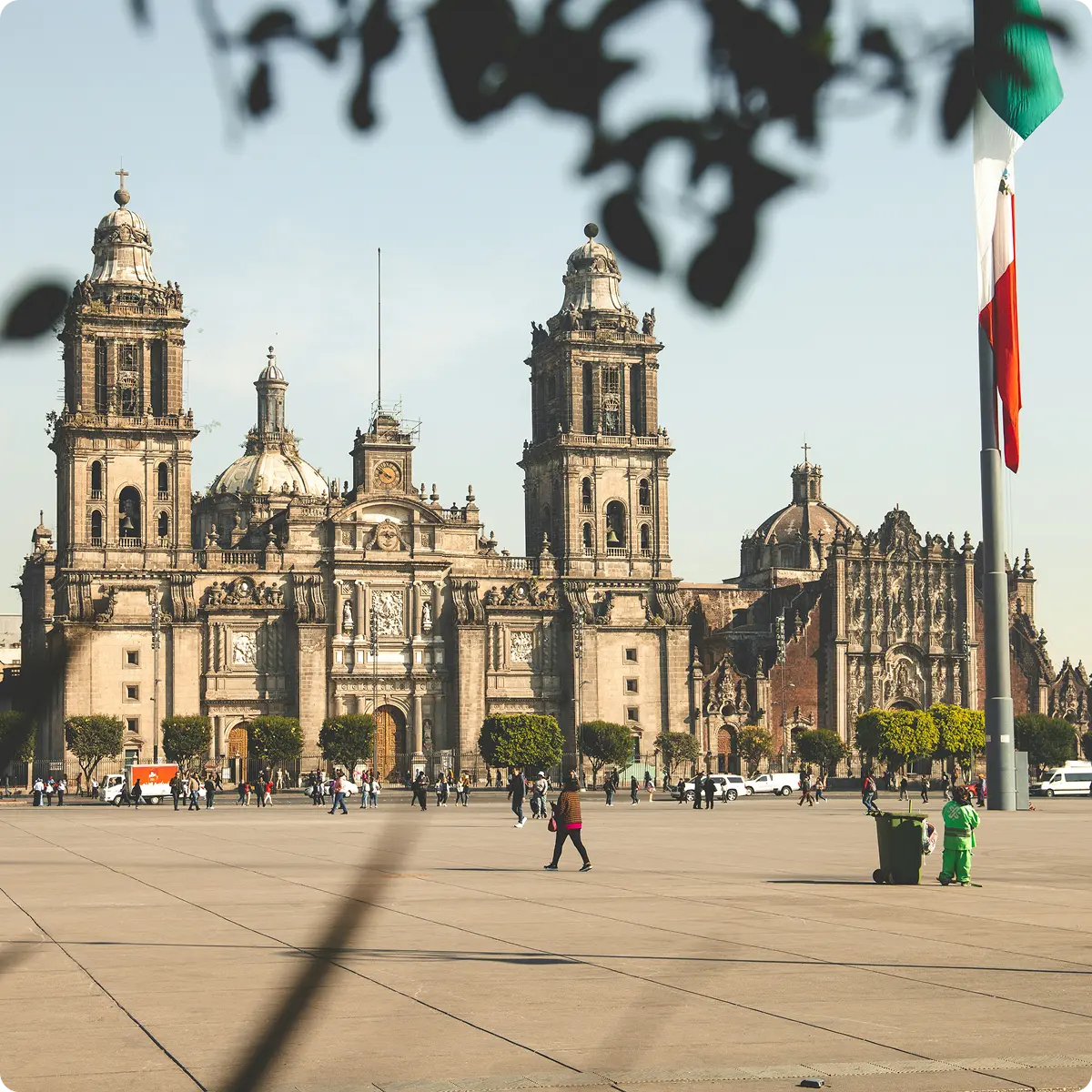 Wide plaza in front of the Mexico City Metropolitan Cathedral with a large Mexican flag and people walking.
