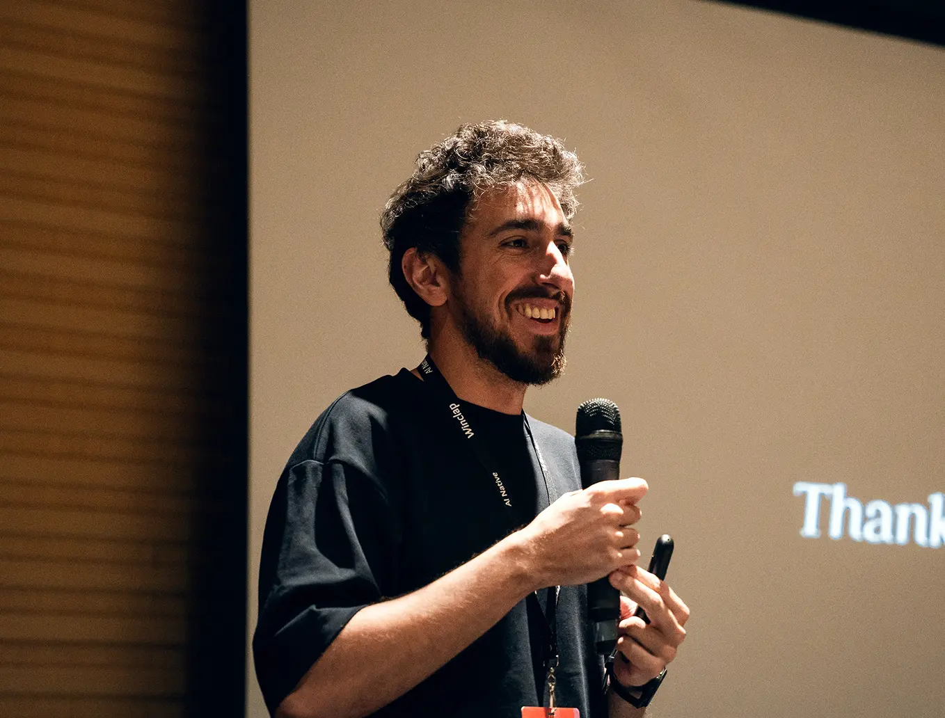 Smiling man with curly hair and beard speaking into a microphone during a presentation.