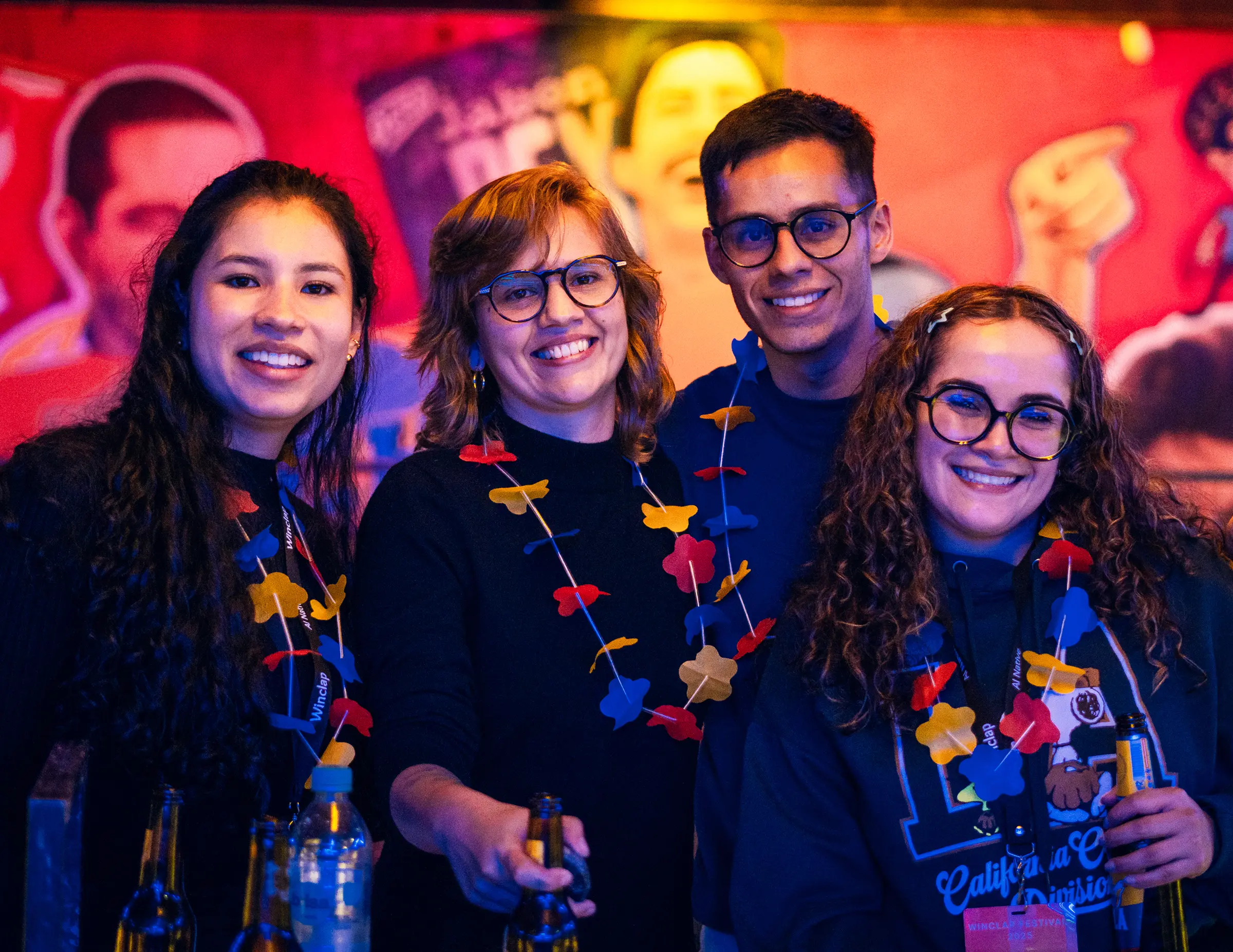Group of four young adults smiling and wearing colorful flower leis at a festive indoor event.