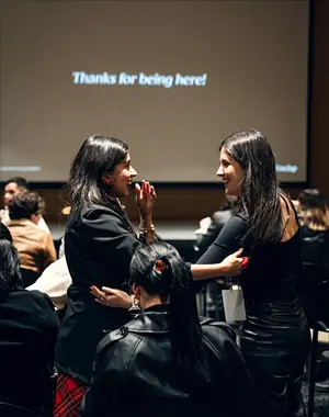 Two women smiling and embracing each other in an audience with a screen showing 'Thanks for being here!' in the background.