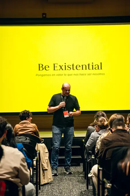 Man holding microphone speaking to an audience with a yellow screen behind him displaying the words 'Be Existential' and a subtitle in Spanish.