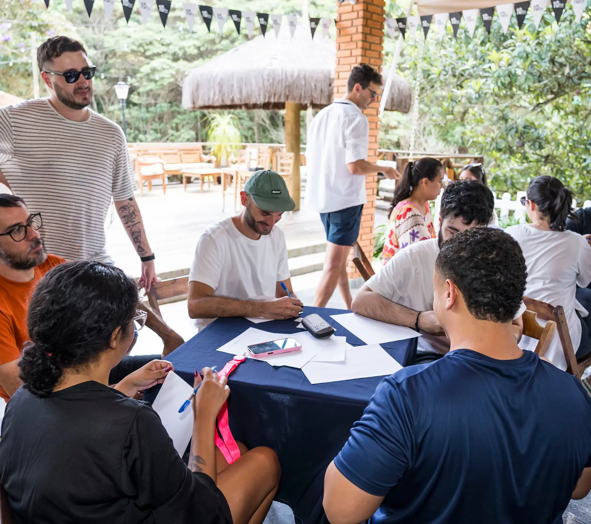 A group of young people sitting around a table outdoors, writing on papers and engaging in discussion under a canopy with greenery in the background.