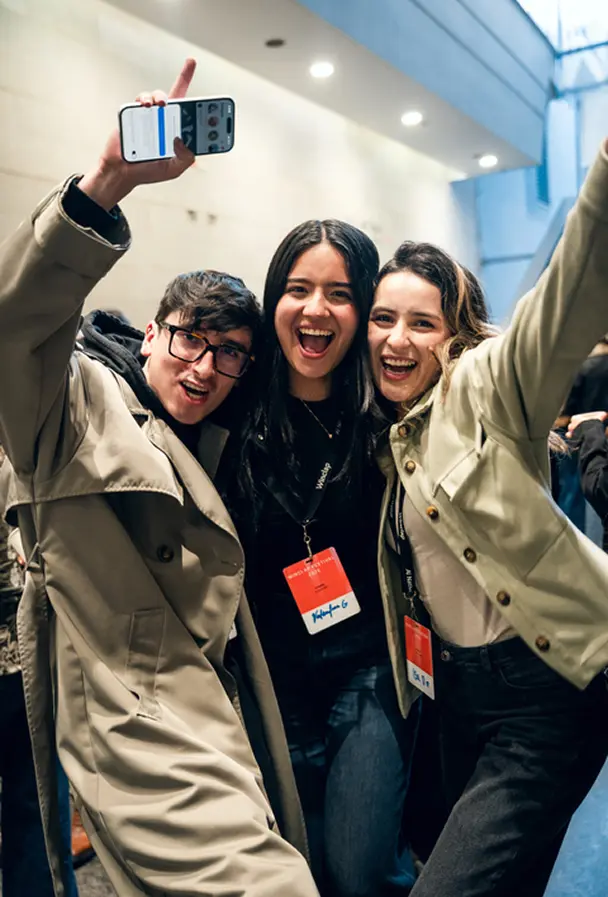 Three young adults smiling and posing excitedly for a selfie in an indoor setting.