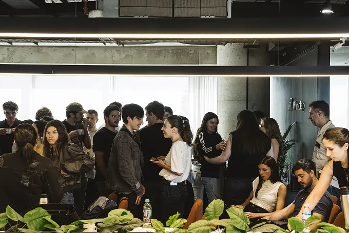 A group of young adults socializing and chatting in a modern office space with indoor plants in foreground.