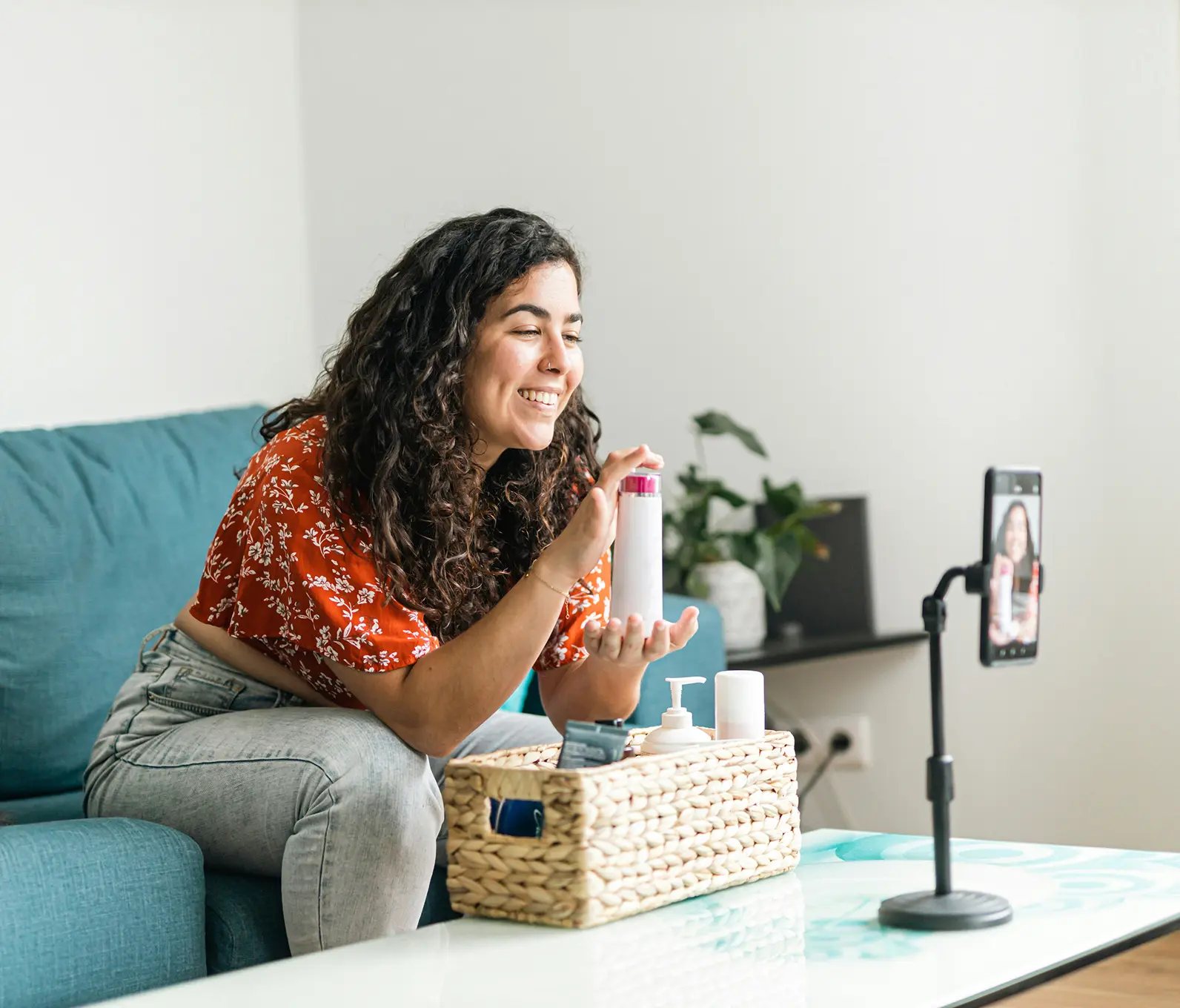 Smiling woman demonstrating a skincare product on video using a smartphone on a stand in a bright living room.