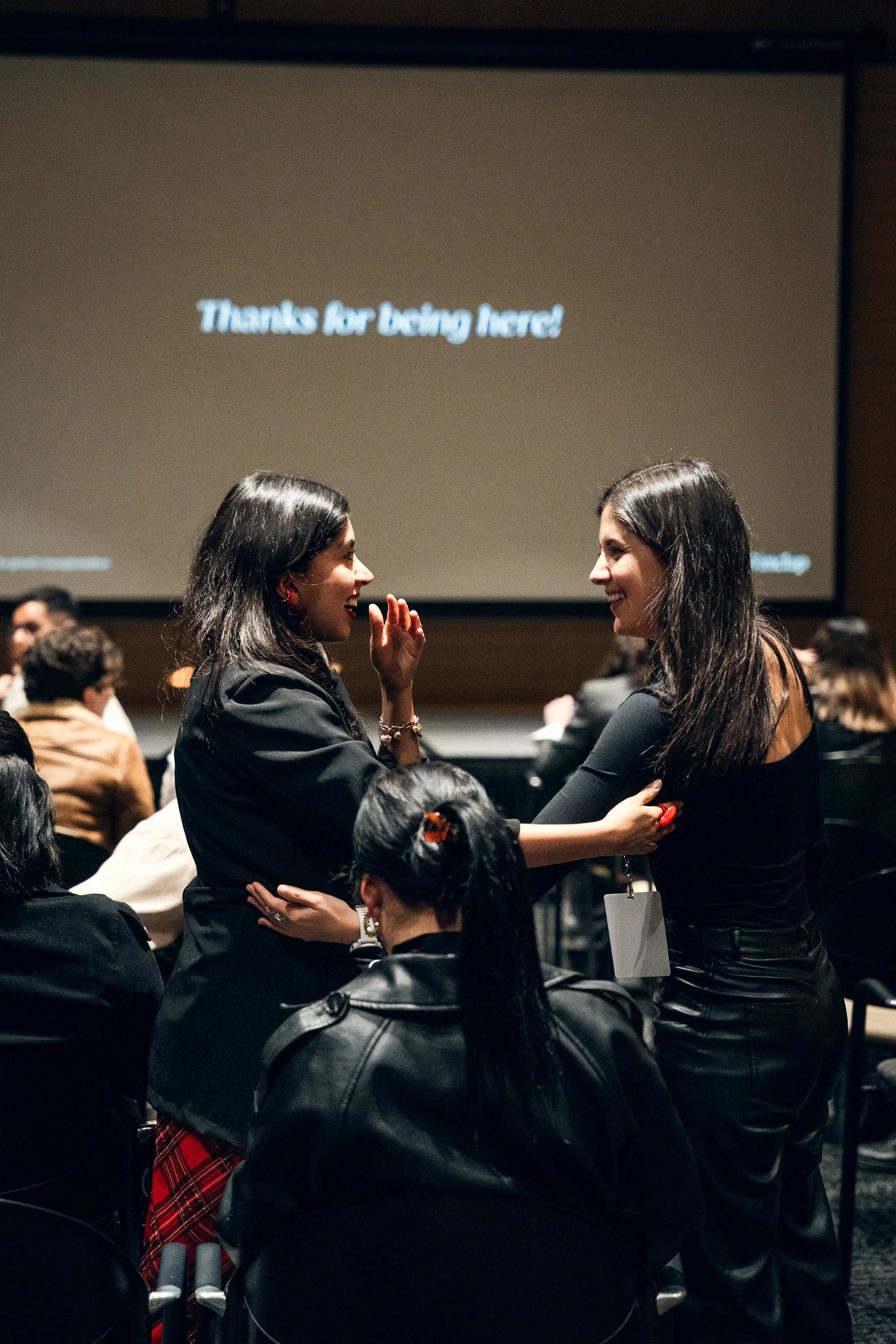 Two women smiling and embracing in a seated audience with a projection screen displaying 'Thanks for being here!' in the background.