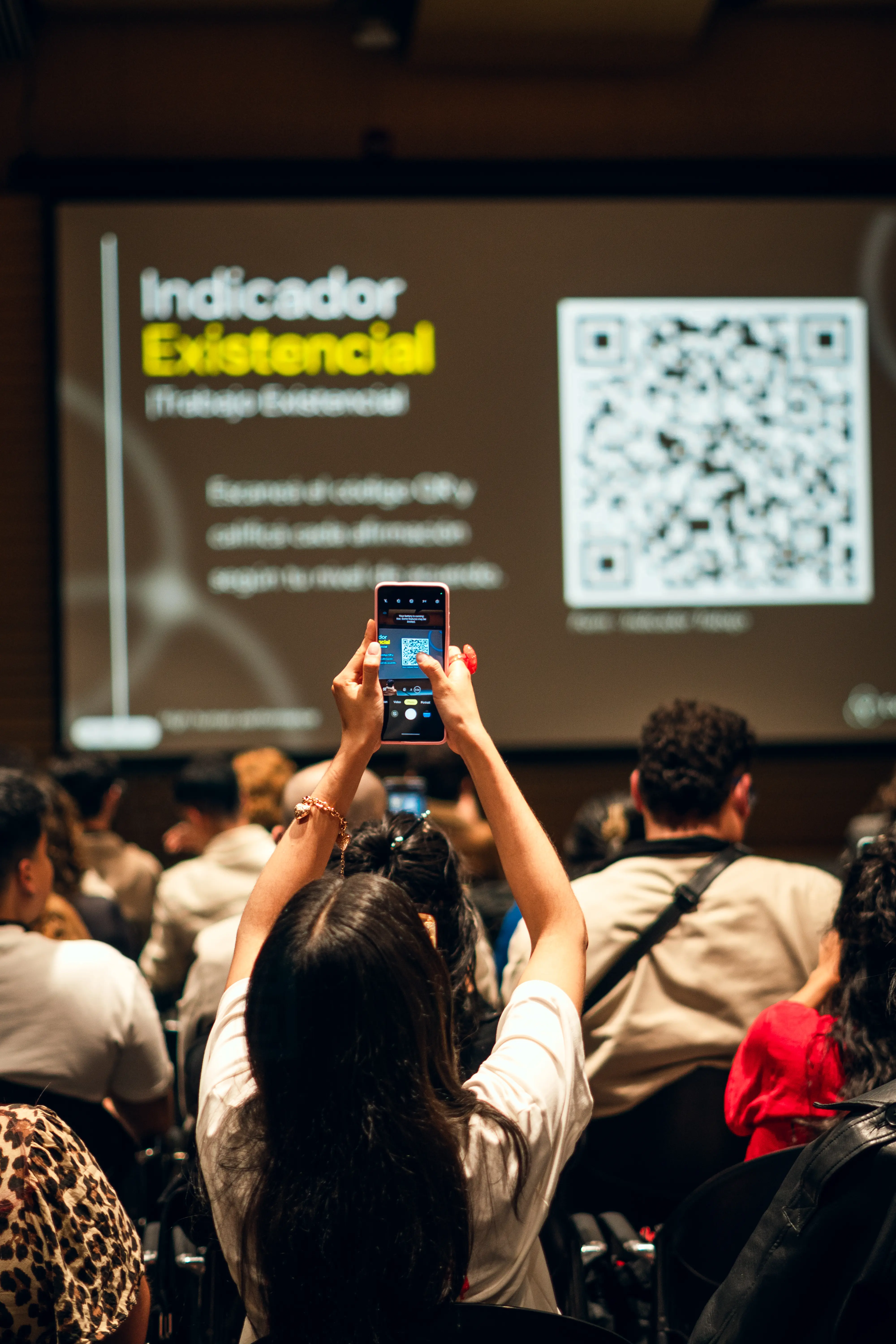 Person in an audience holds up a phone to scan a QR code displayed on a presentation screen.