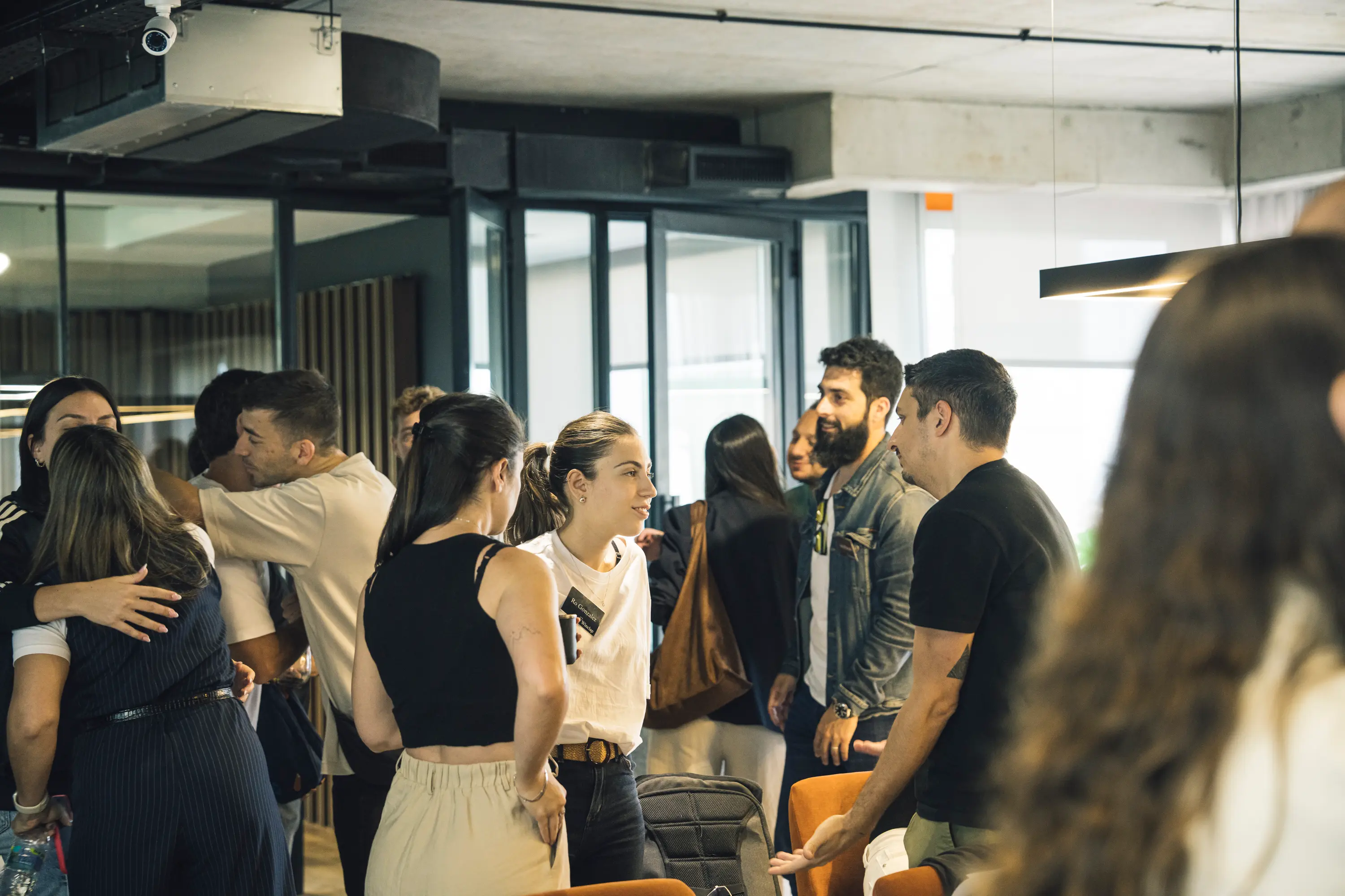 Group of young adults socializing and greeting each other in a modern indoor space with glass doors and orange chairs.