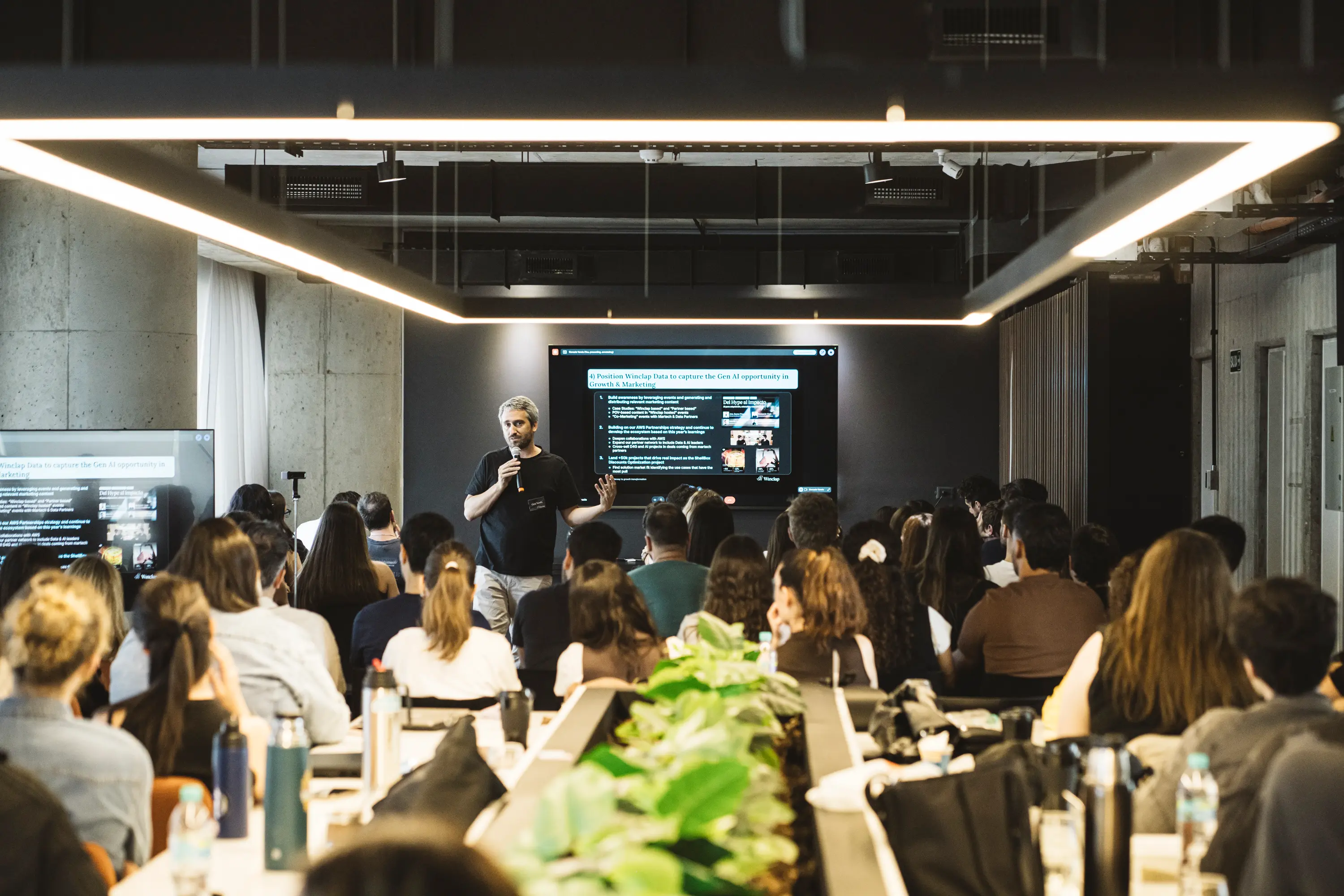 Man giving a presentation to a seated audience in a modern conference room with two screens displaying slides.