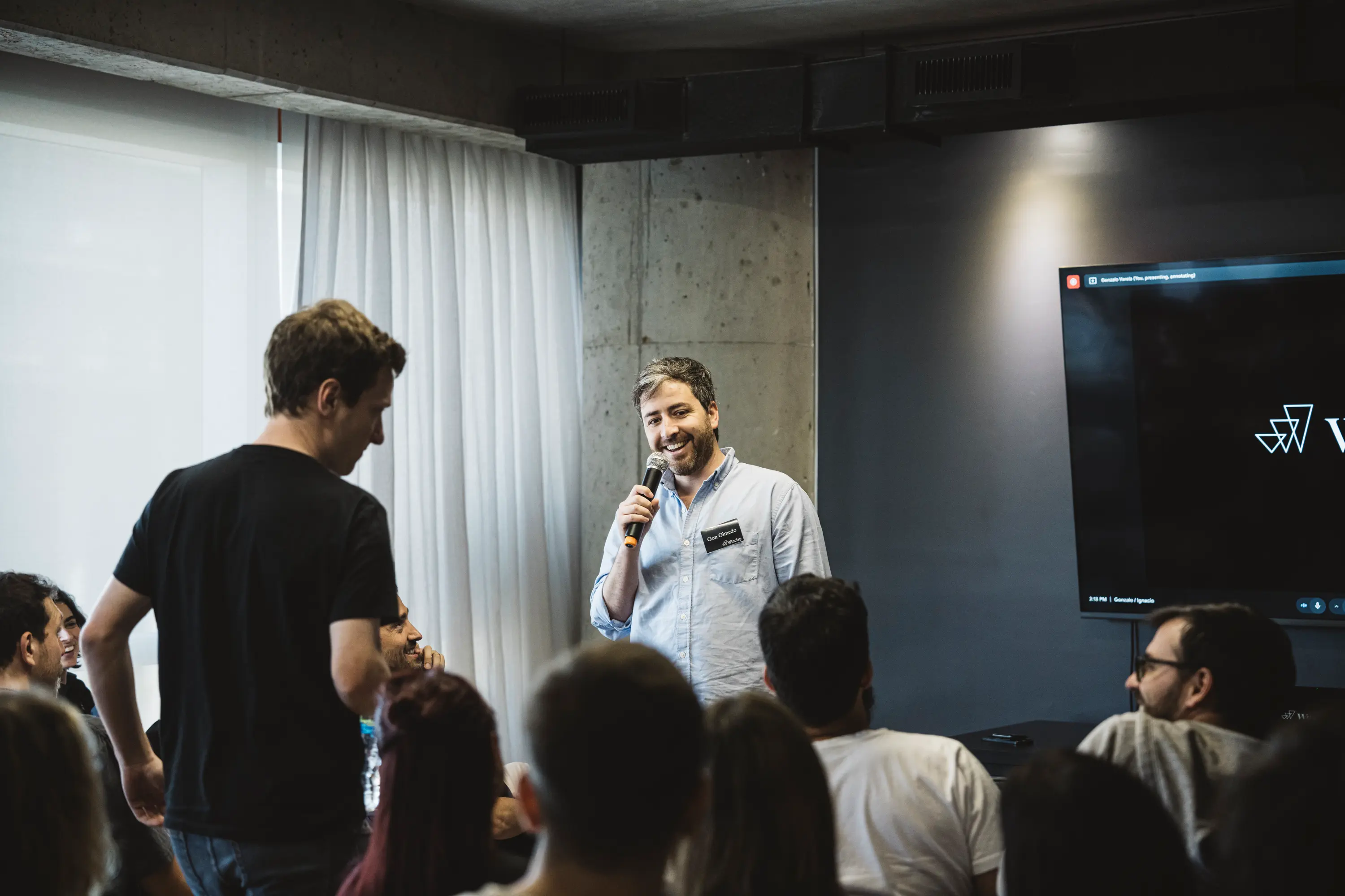 Man holding a microphone speaking to an attentive audience in a modern indoor setting.