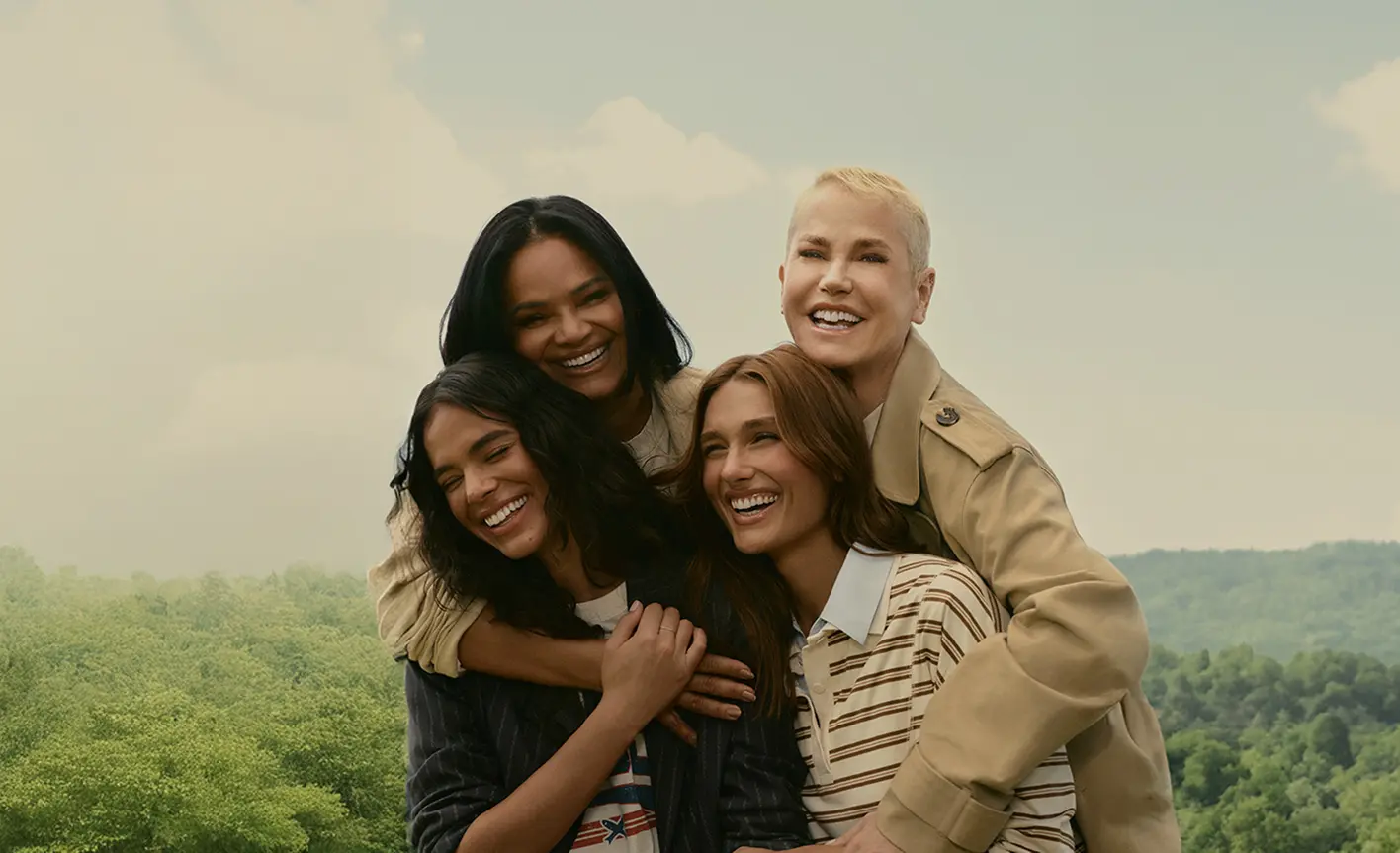 Four women smiling and hugging each other outdoors with green trees and a cloudy sky in the background.