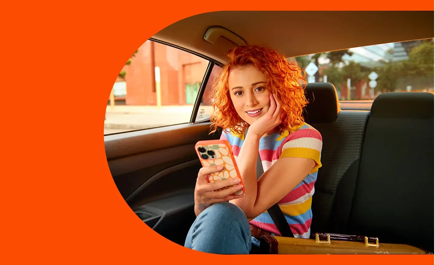 Young woman with curly red hair sitting in the backseat of a car, smiling while looking at her phone.